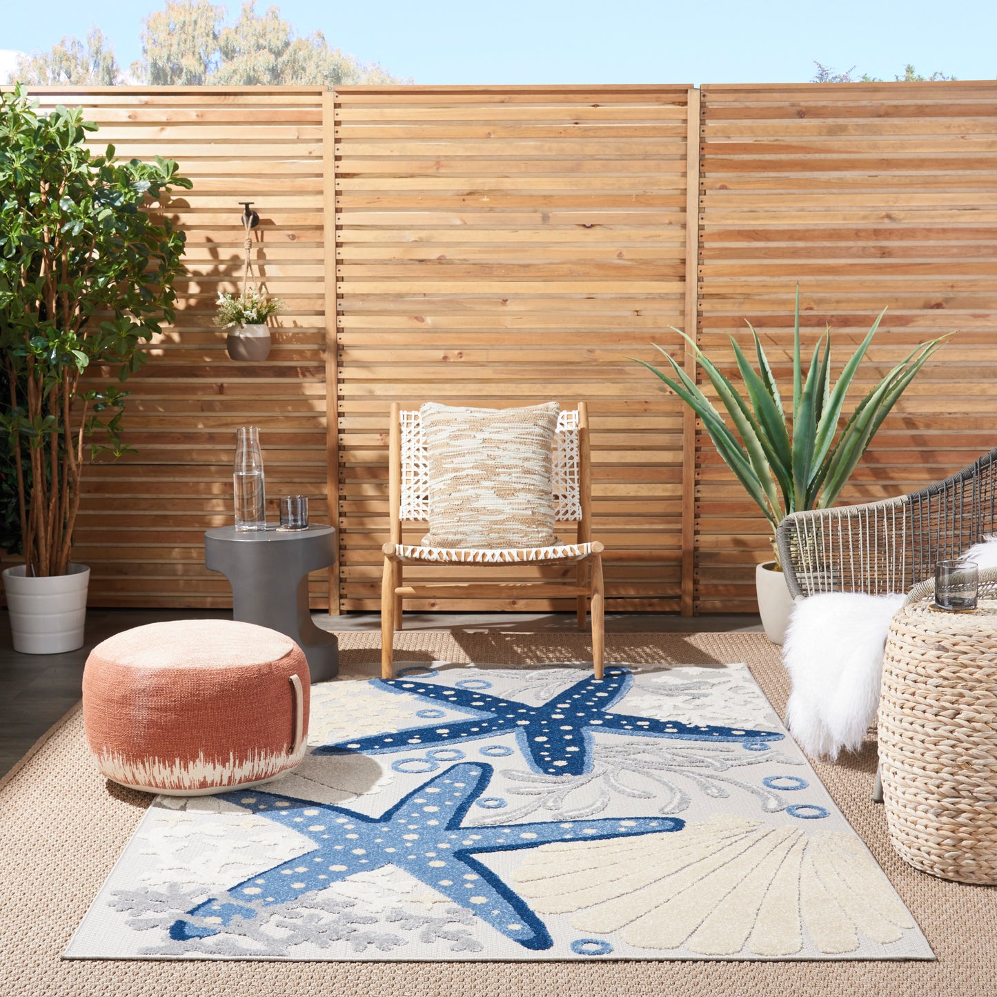 Outdoor patio with wooden slat fence, woven chair, pink pouf, blue starfish rug, and potted plants.