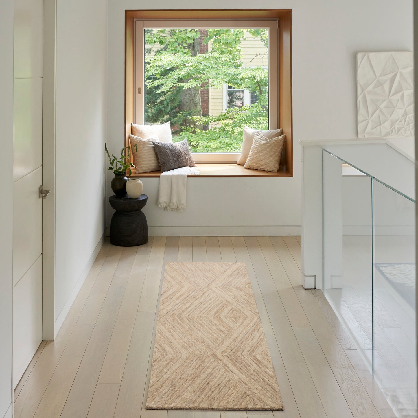 Bright minimalist hallway with wood-framed window seat, beige runner rug, cushions, plant on a black side table, glass railing.