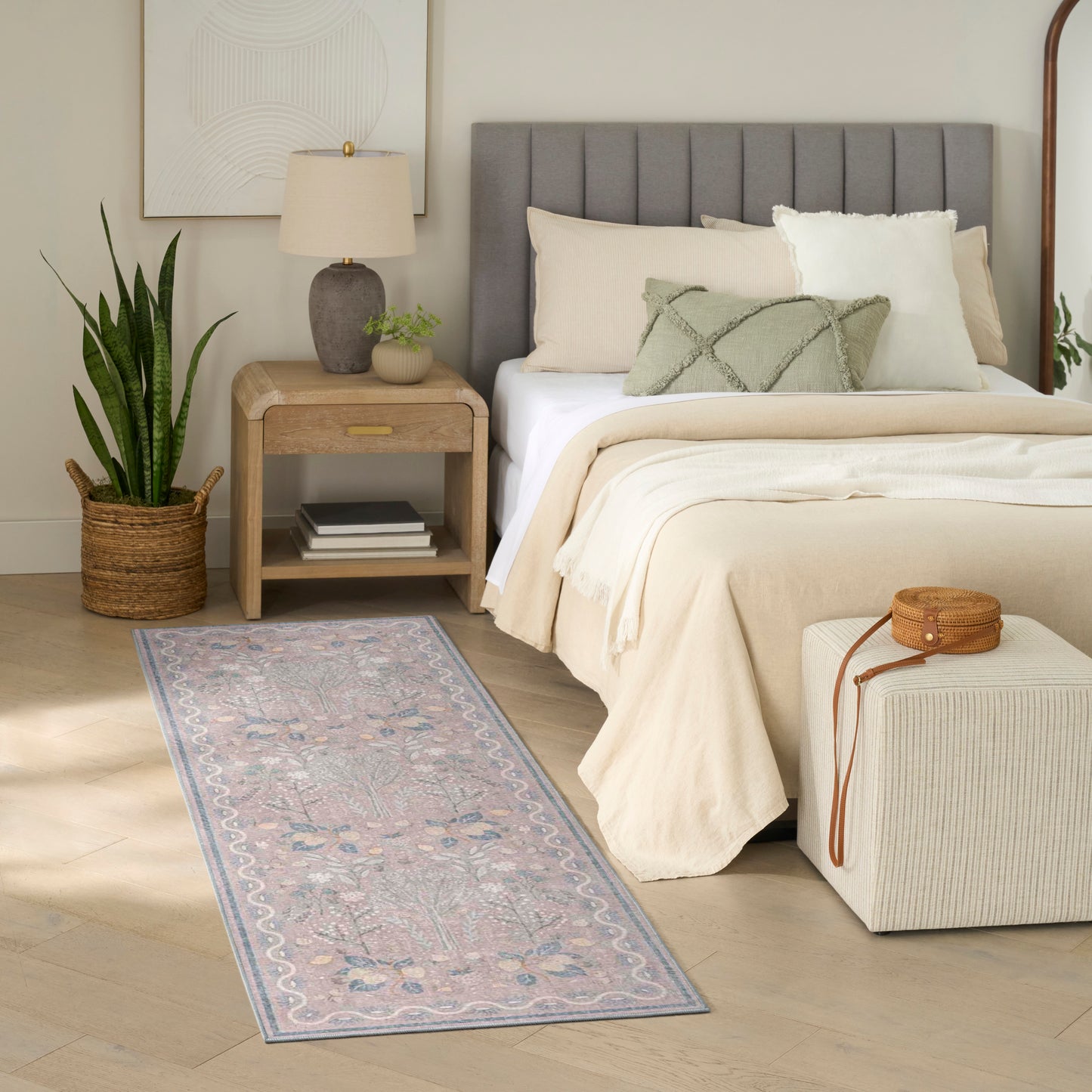Neutral bedroom with beige bedding, gray tufted headboard, wooden nightstand, snake plant, and pink floral runner rug.