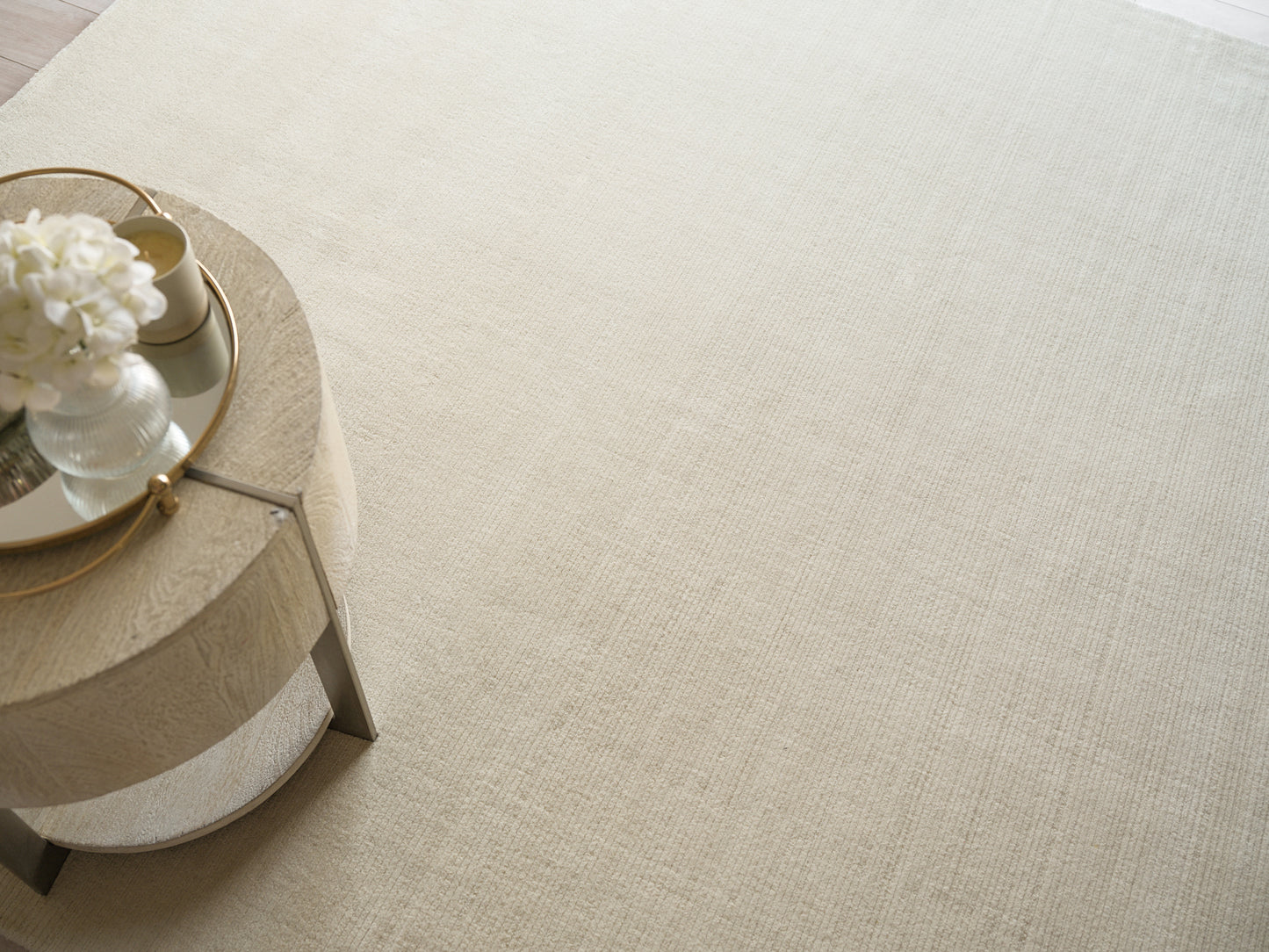 Round glass-top side table with gold metal frame on a light beige textured rug, vase of white flowers.
