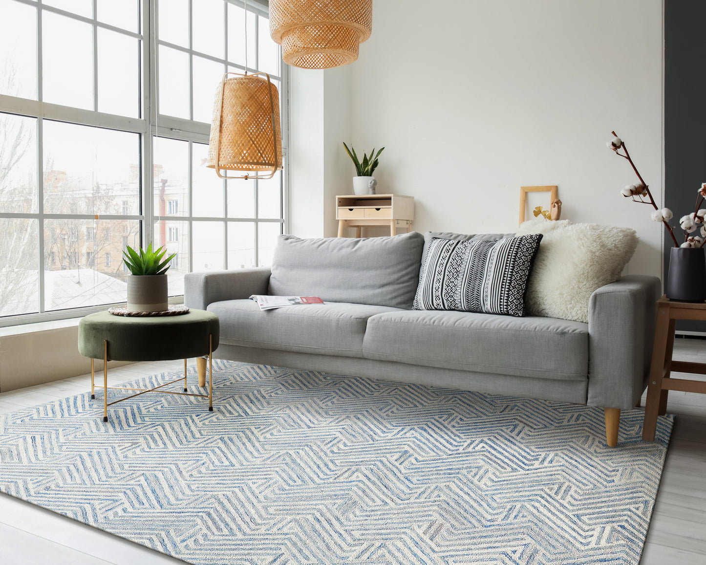 Light gray sofa with patterned cushions in a bright living room; blue-white chevron rug, wicker pendant lights, plants.