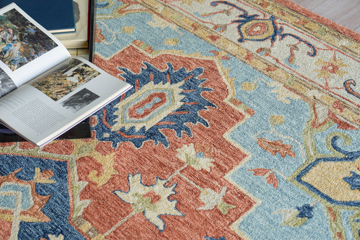 Vibrant Persian-style area rug with red, blue, gold tones & floral motifs; open magazine & books rest on edge.