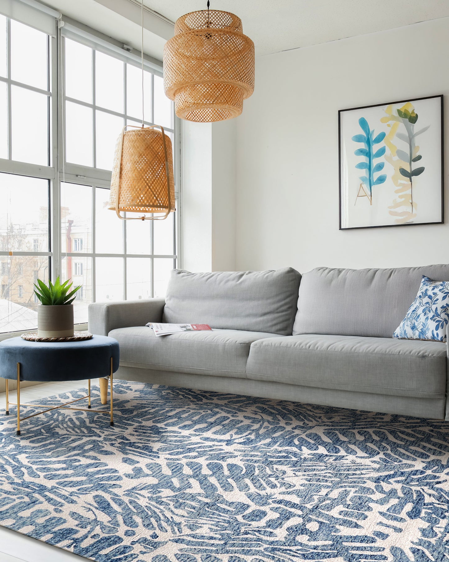 Modern living room with gray sectional, blue leaf-pattern rug, wicker pendant lamps, blue ottoman and botanical wall art.