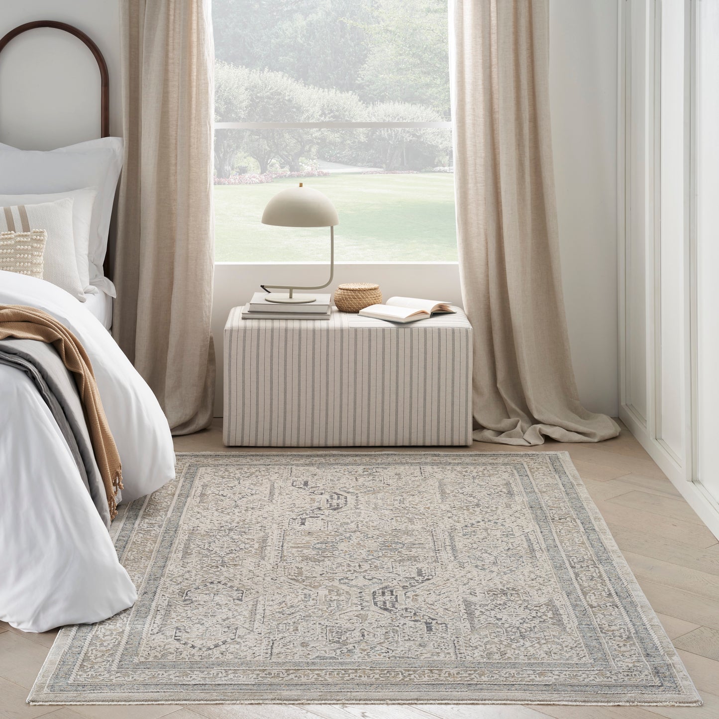 Light gray-cream patterned area rug with a medallion border in a sunlit neutral bedroom beside a striped rectangular ottoman.