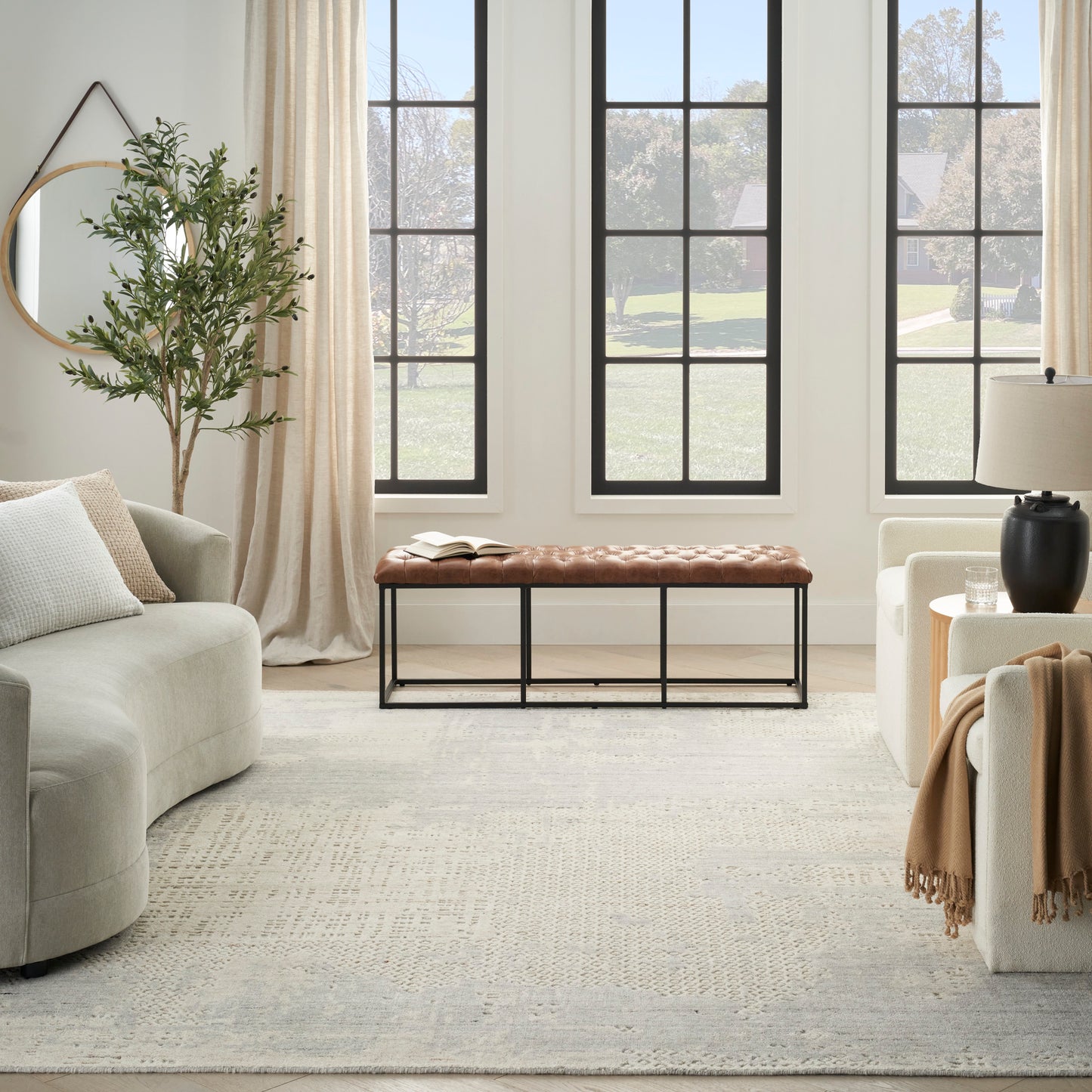 Long brown tufted leather bench with black metal frame in a bright neutral living room with large grid windows.