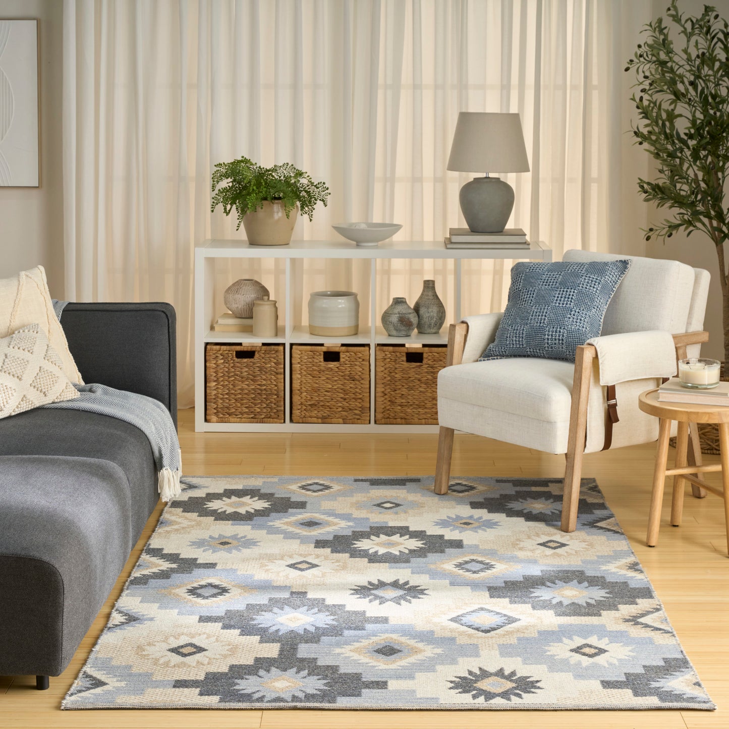 Cozy living room with light curtains, white shelves, baskets, gray sofa, cream armchair, blue pillow, and a blue-gray rug.