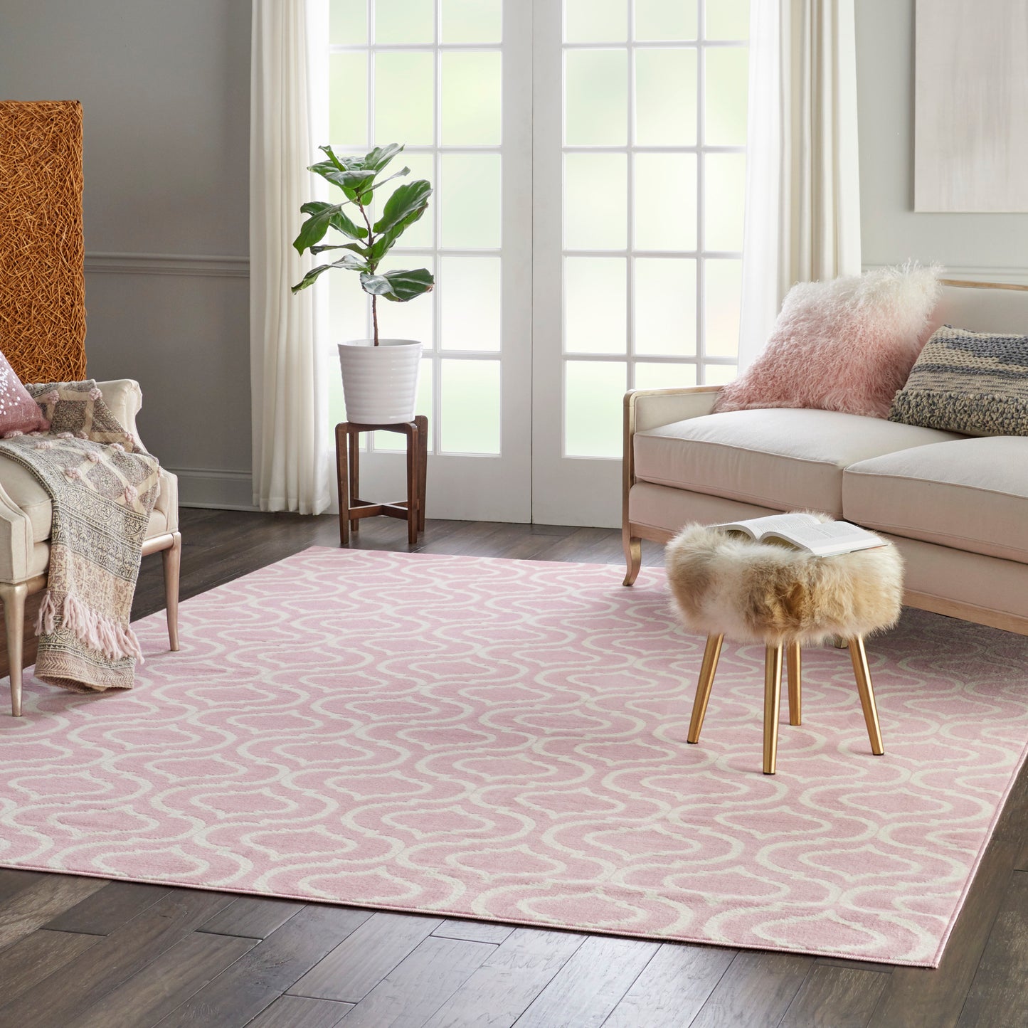 Soft pink geometric area rug in a bright living room with a cream sofa, fur stool, potted plant, and large windows.
