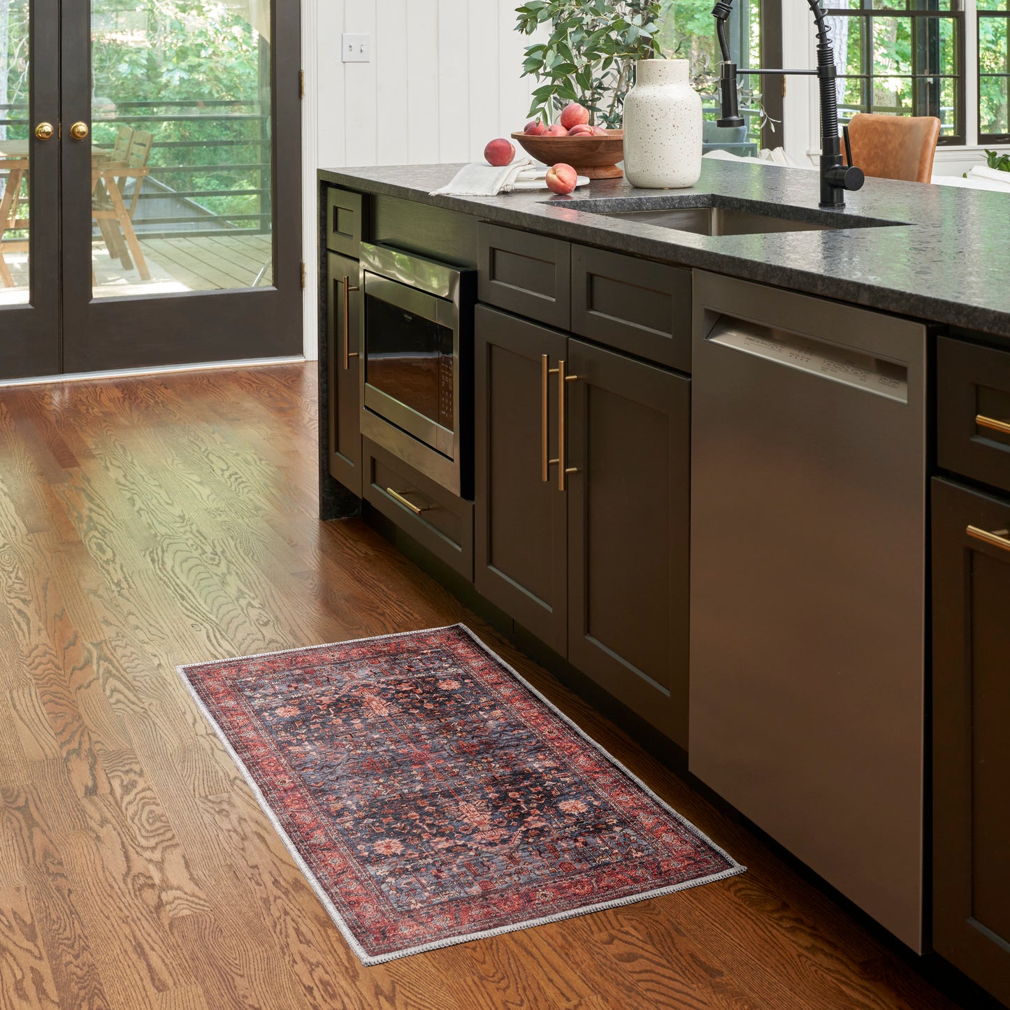Dark wood kitchen with stainless dishwasher, built-in oven, granite counter, hardwood floors, red rug runner, vase and plants.