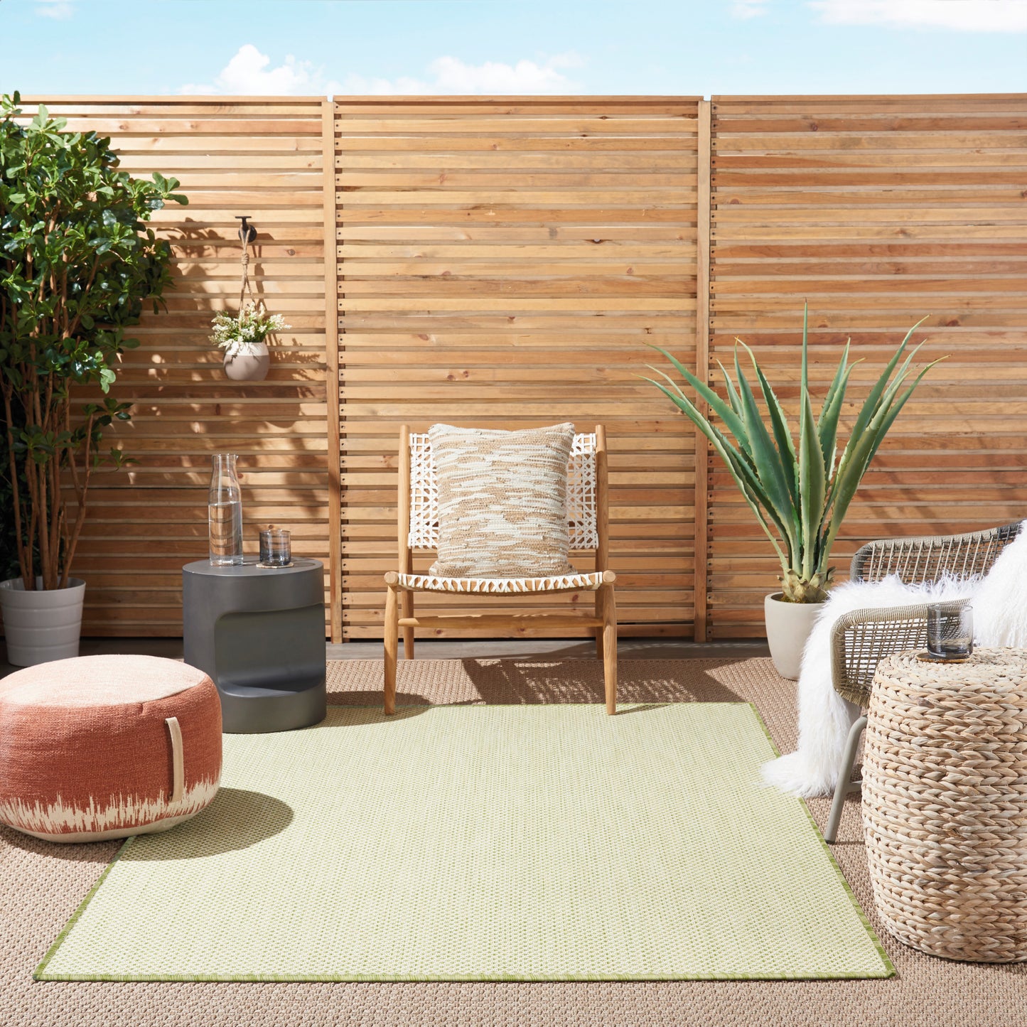 Boho outdoor patio with wooden slat fence, woven armchair w/ cream cushion, orange pouf, pale green rug, and potted plants.