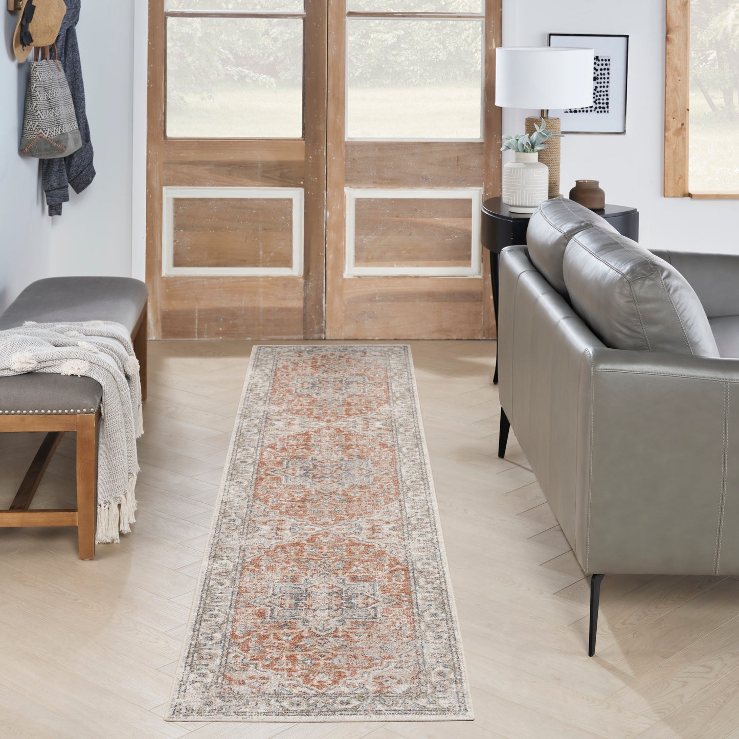 Rustic living room entry with a vintage 8' runner rug in orange and grey, a grey leather sofa, wooden double doors, and a bench with a throw.