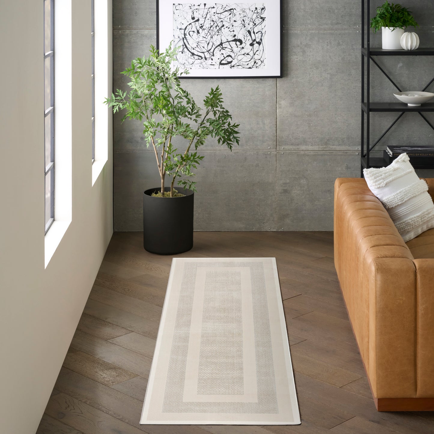 Light-gray runner rug with double border beside a tan leather sofa, green potted plant & black metal shelf.