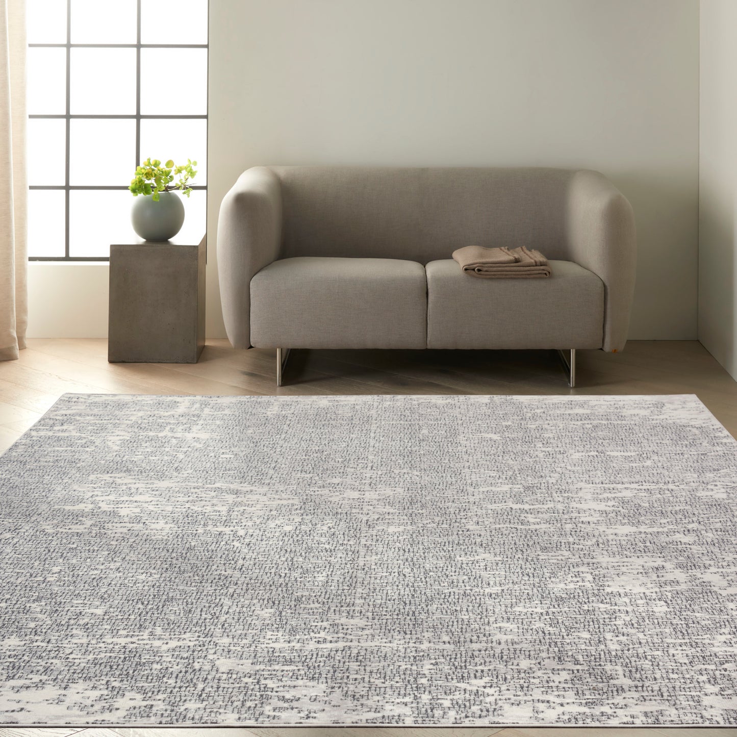 Light gray modern loveseat with rounded arms on metal legs, beside a concrete side table and plant on a gray patterned rug.