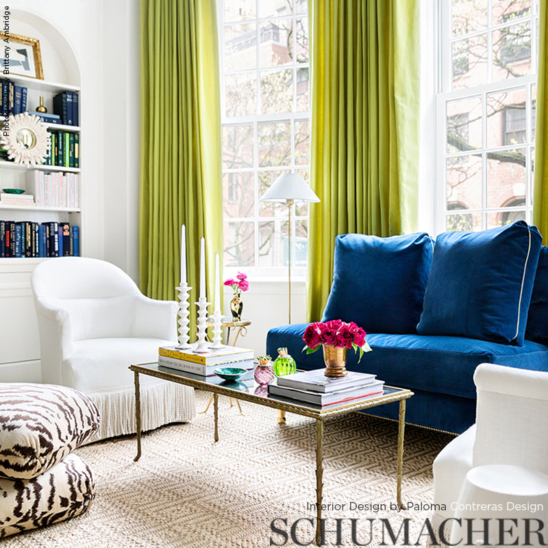Sunny living room with lime-green curtains, blue velvet sofa, white armchairs, glass coffee table, zebra-print ottoman, and bookshelves.