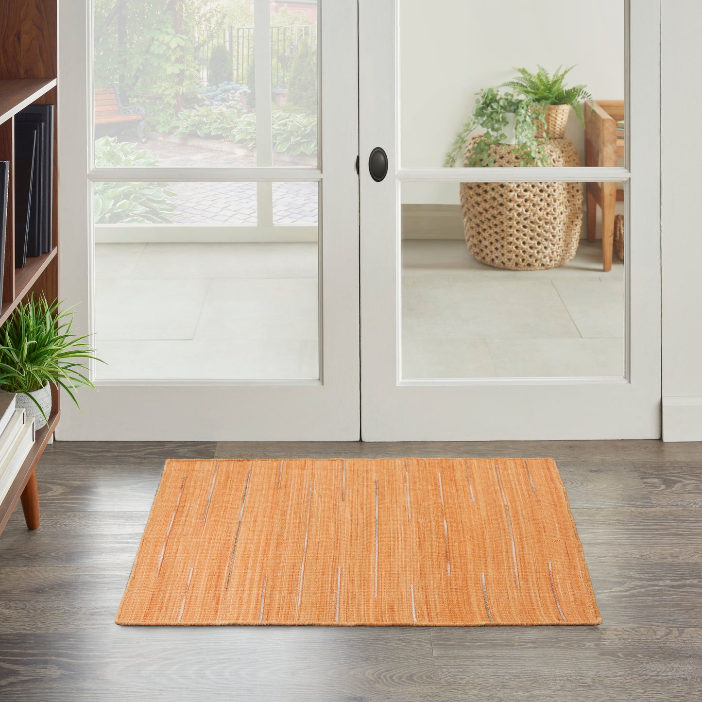 Rectangular orange flat-weave rug in front of glass-paned white doors & dark wood floor; nearby houseplants.