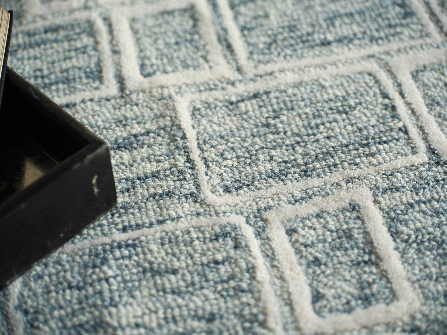 Close-up of a blue woven rug with white geometric rectangle patterns; corner of a black box visible.