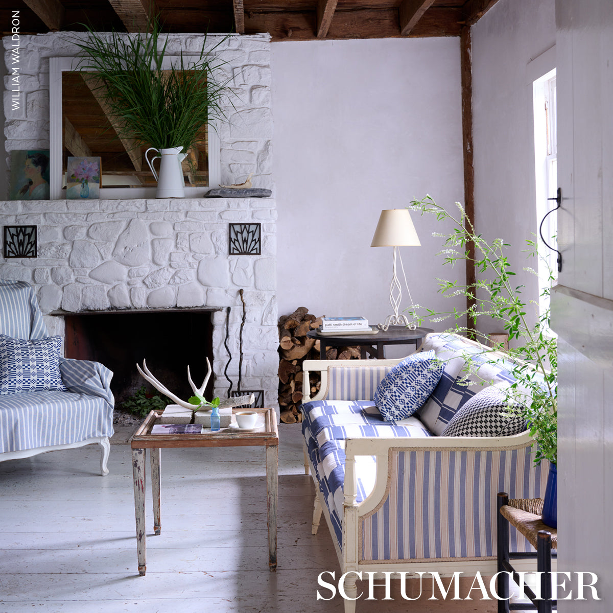 Cozy living room with white stone fireplace, blue-striped sofas, wood coffee table, potted plants, stacked firewood, lamp.