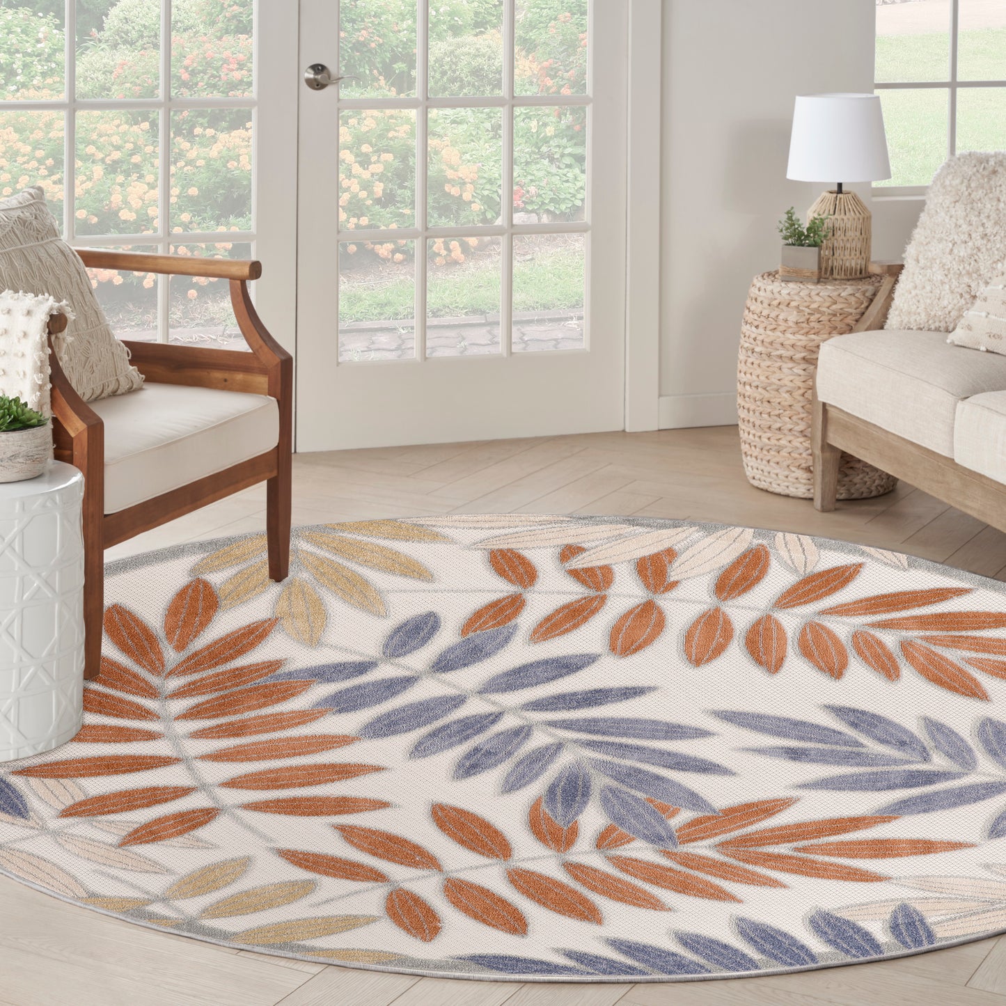 Round woven rug with orange, blue, and tan leaf pattern on a light cream background in a sunlit living room.