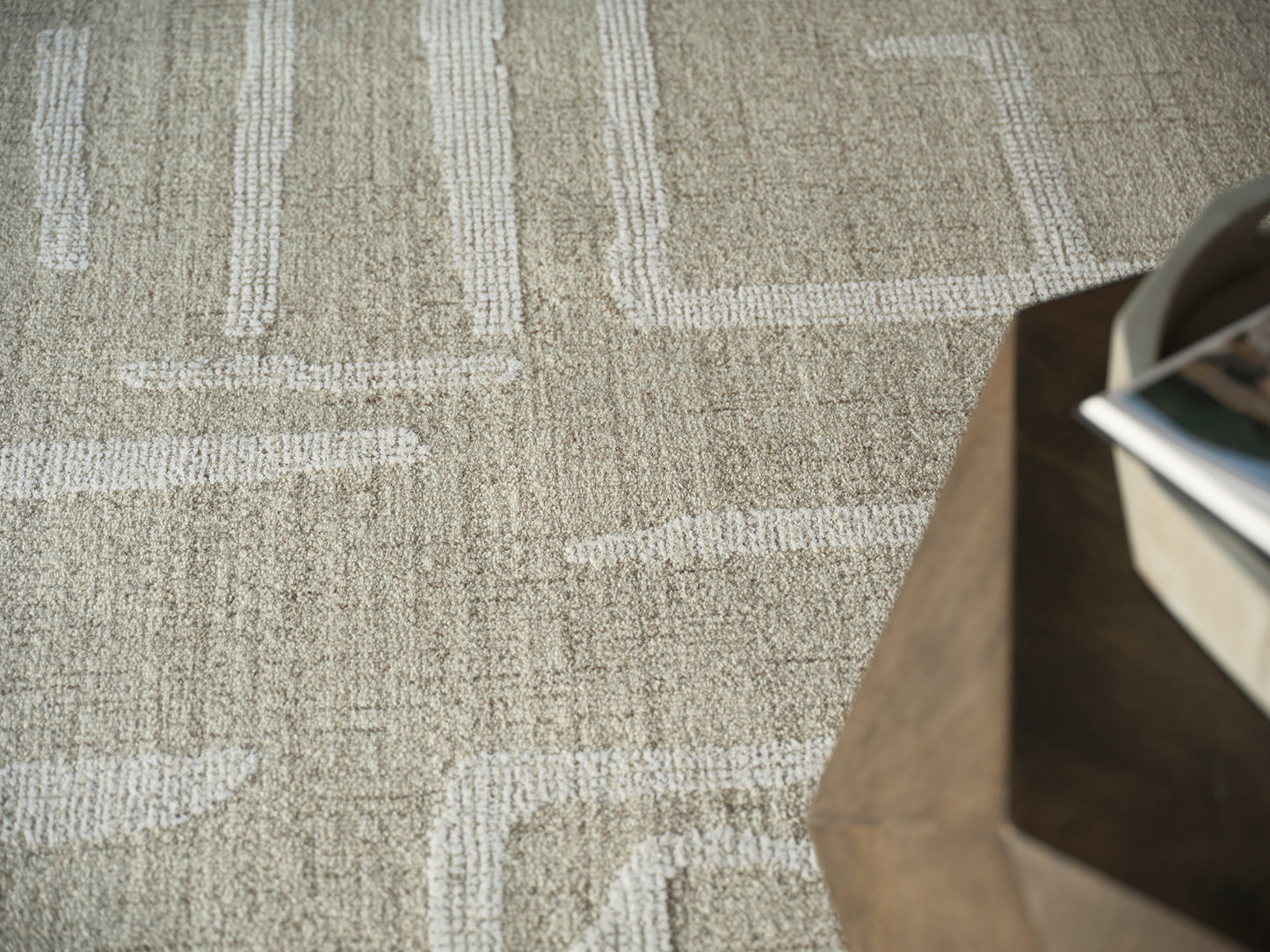 Beige textured rug with white geometric lines; dark wooden table edge on the right with magazines.