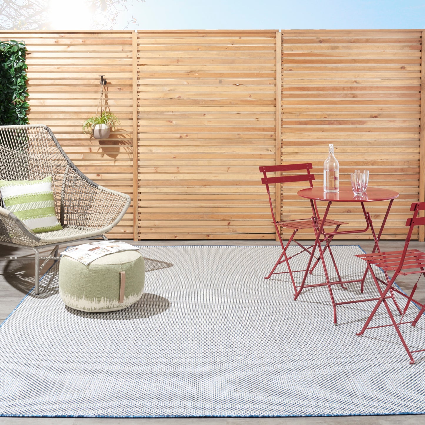 Outdoor patio with wicker chair, green cushion, round pouf, red metal bistro set, light rug, and wooden privacy fence.