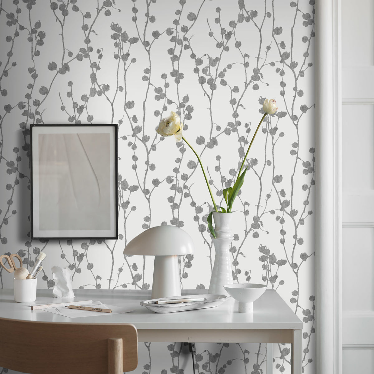 White desk with a modern lamp, vase of white lilies, framed art, light wood chair, and grey vine wallpaper.