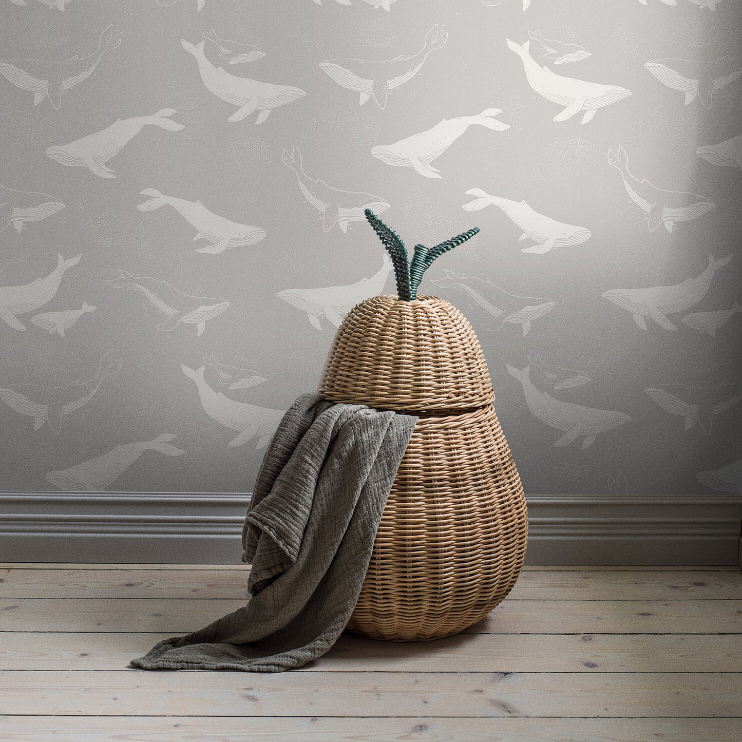 Natural woven pear-shaped basket with lid, green stems, draped blanket, light wood floor, gray wallpaper with white birds.