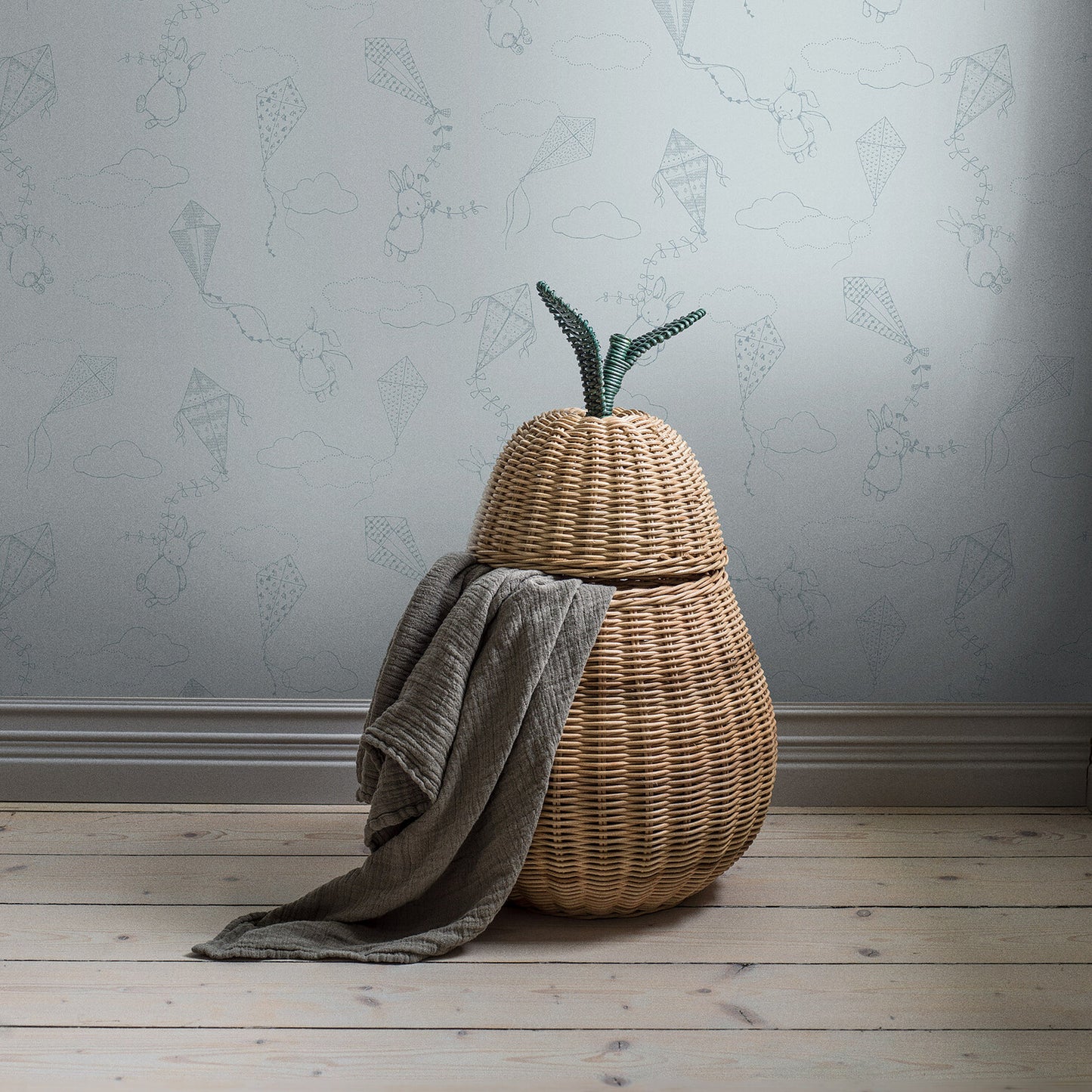 Pear-shaped woven rattan basket with lid and green stem, gray knit blanket draped over it, light wood floor.
