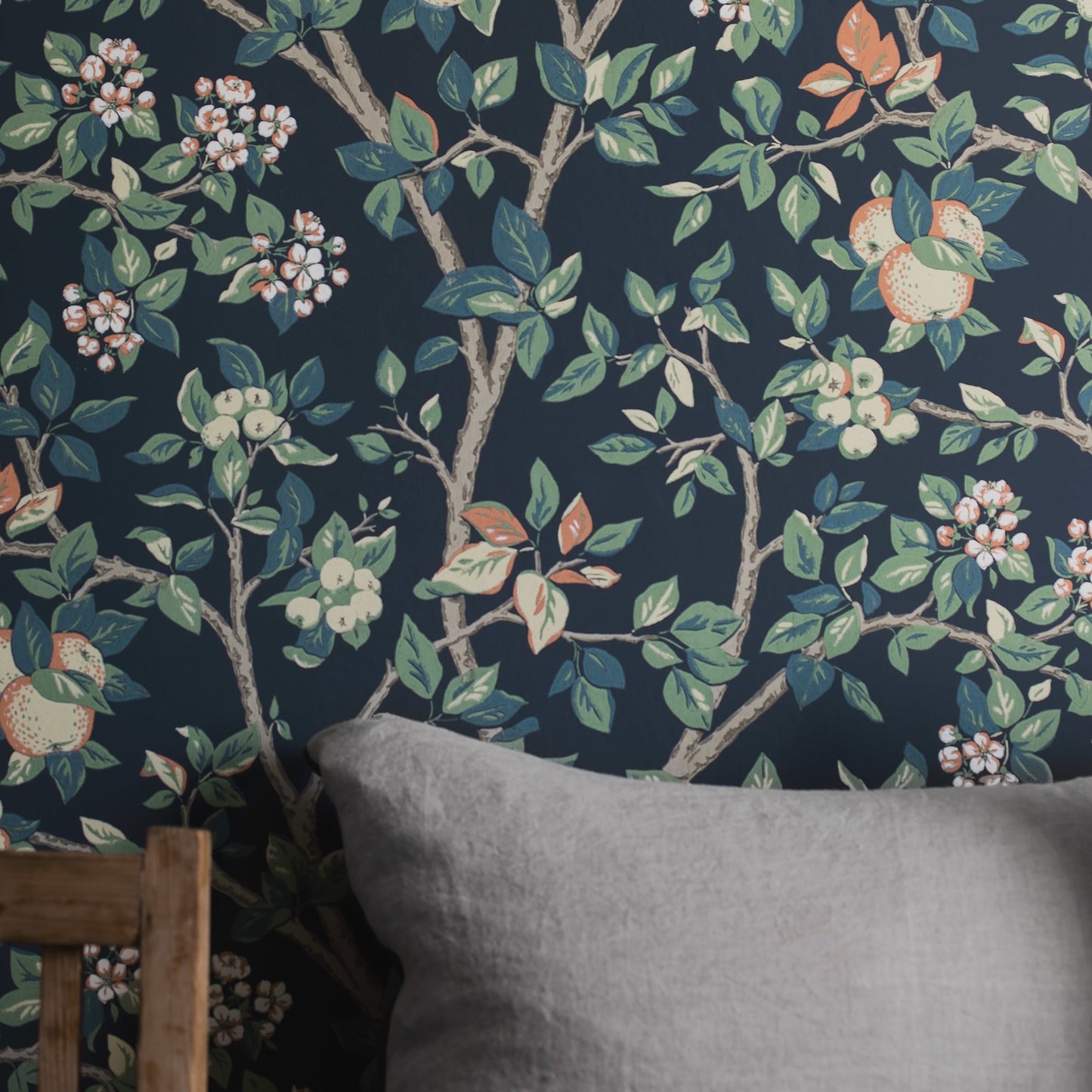 Botanical wallpaper: navy background with brown branches, green leaves & pink blossoms with fruit; shown with a gray cushion.