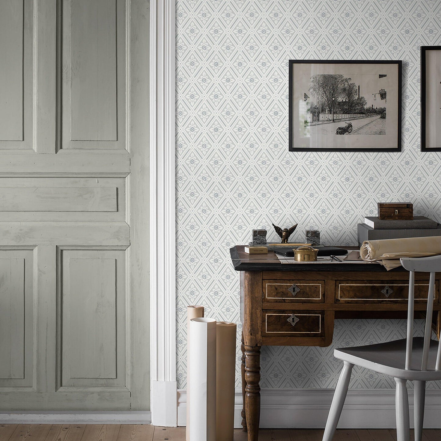 Patterned neutral wallpaper with mineral tones, accompanied by a vintage dark-wood desk, brass hardware, papers, ink, a scroll, and a gray chair.