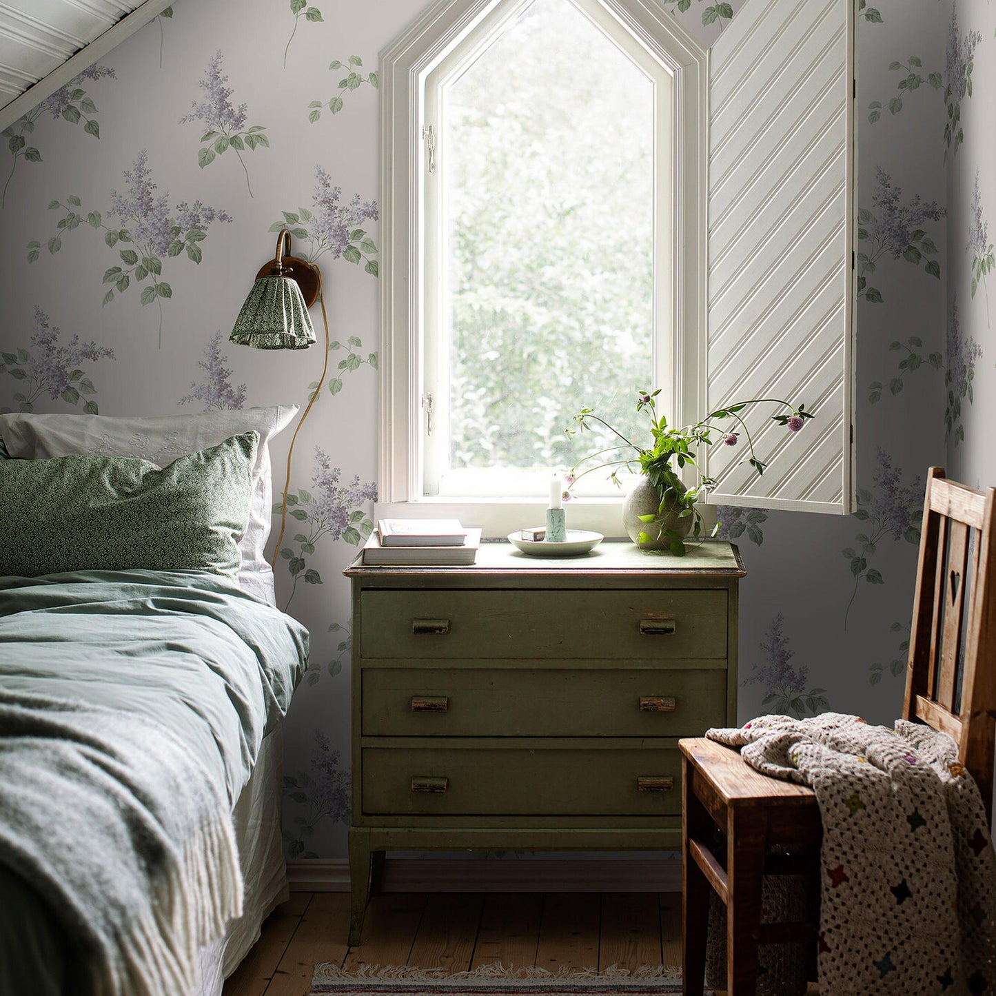 Cozy vintage bedroom with arched window, sage-green dresser, lavender floral wallpaper, bed linens, plant, and wooden chair.
