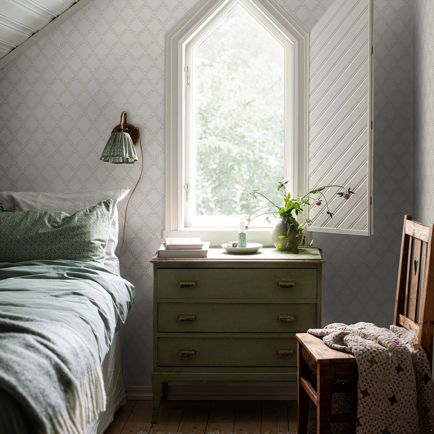 Bedroom with sage green chest, arch window, pale wallpaper, bed with green bedding, wooden chair, vase and books on dresser.