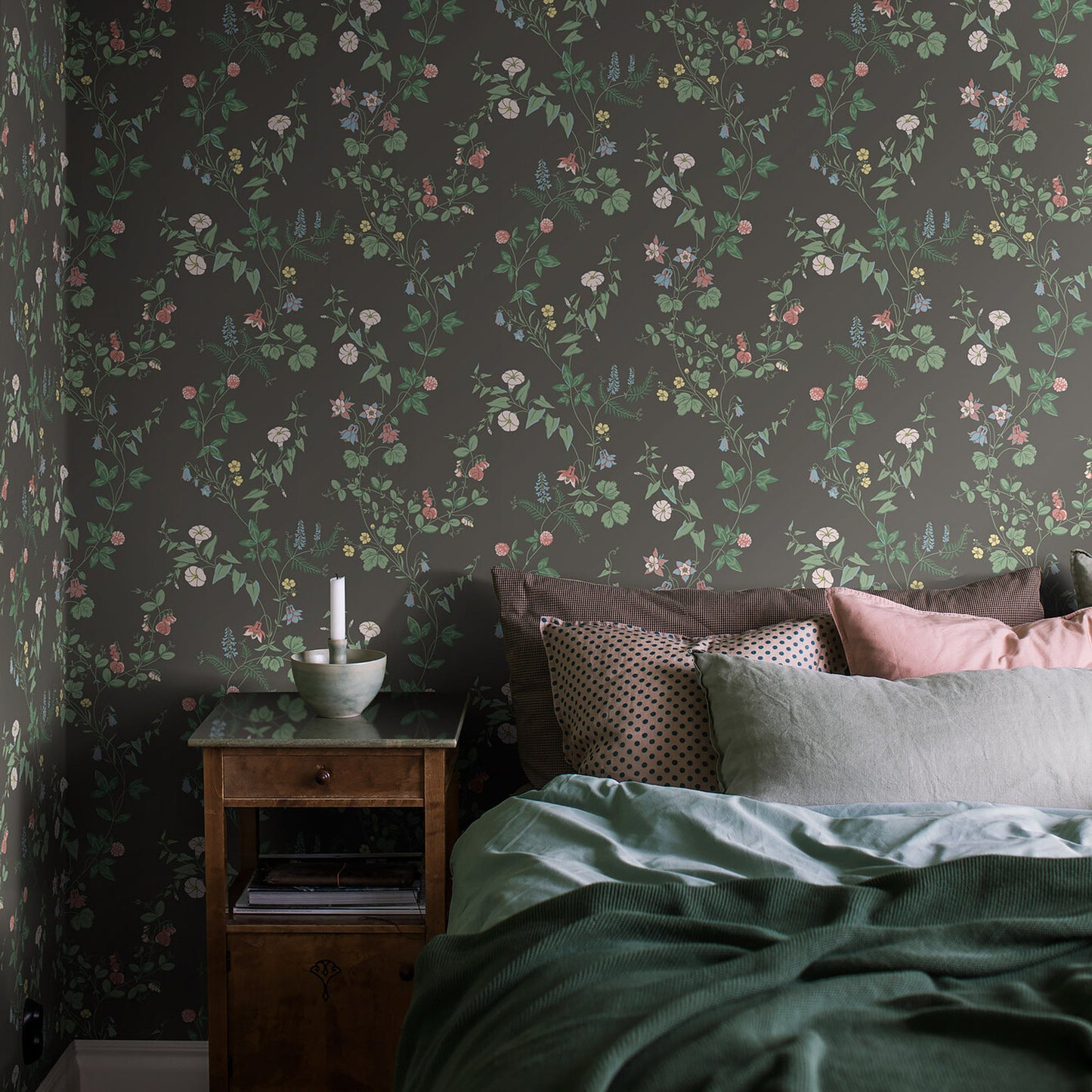 Bedroom with dark botanical wallpaper, wooden nightstand, candle, books, and bed with green duvet and pink/gray pillows.