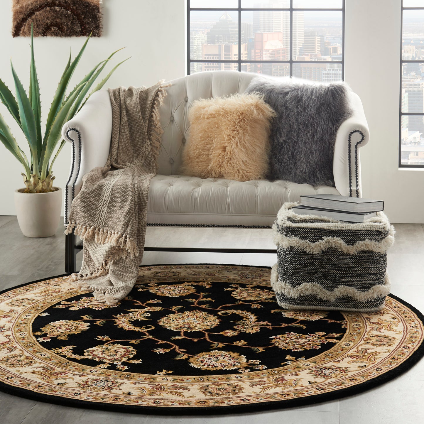 White tufted sofa with beige and gray pillows and throws, round black-gold ornate rug, potted plant, book-stacked ottoman.