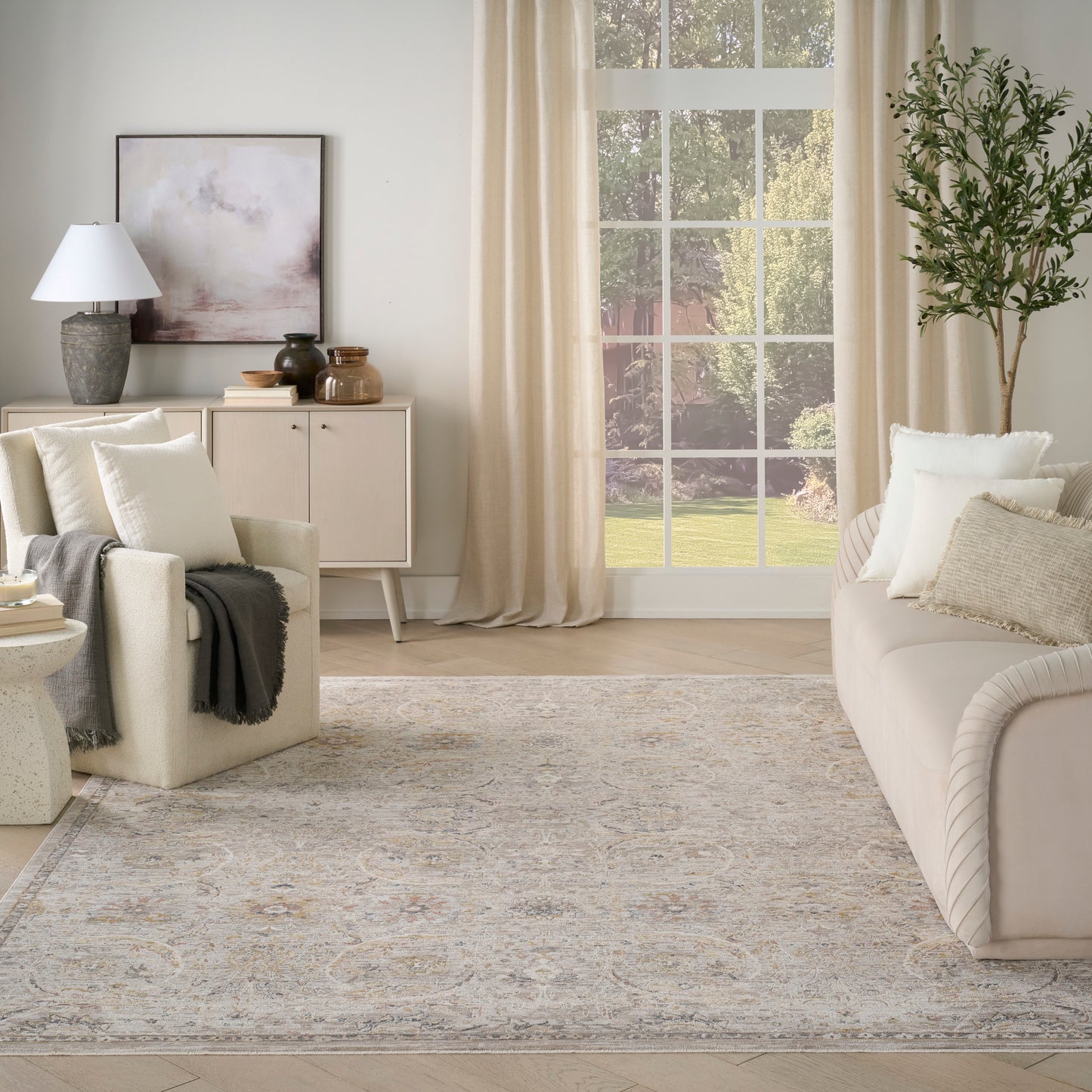Neutral living room with beige fabric sofa, soft throw, light patterned rug, cream curtains, large window, and potted tree.