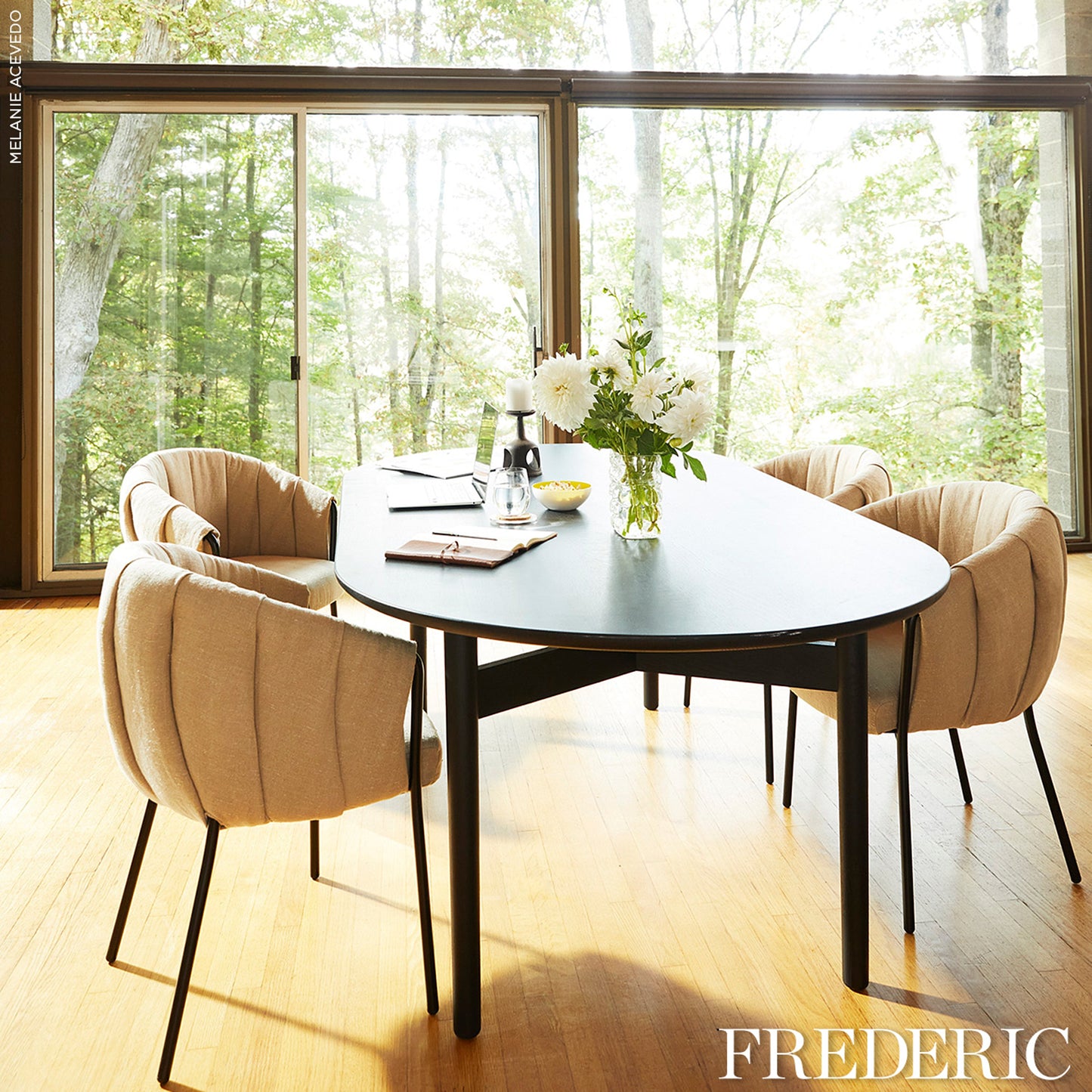 Dark oval dining table with black legs and beige tufted chairs, white flower centerpiece in sunlit forest-view dining room.