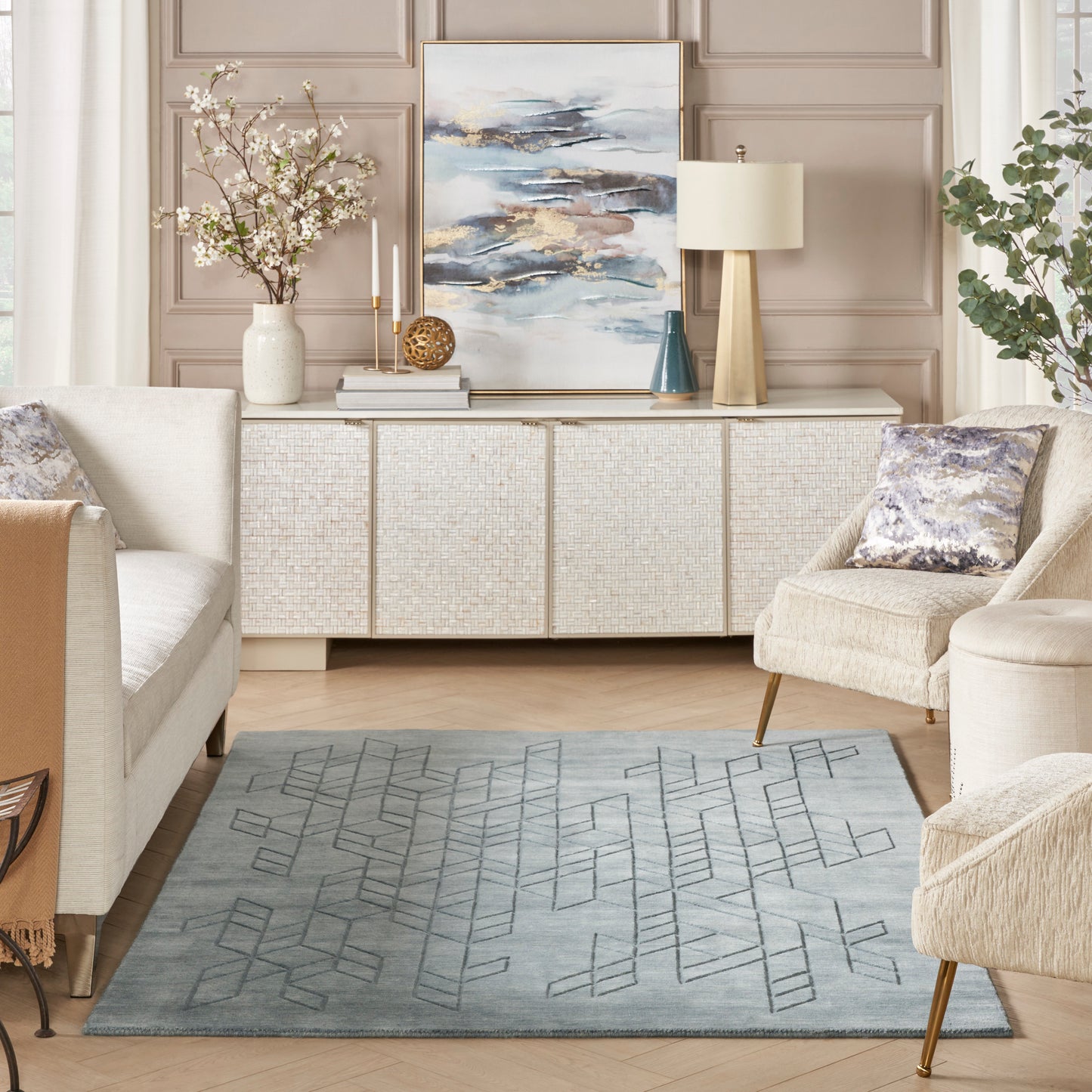 Neutral living room with beige sofa and chair, woven sideboard, abstract blue artwork, vase, and a geometric blue rug.