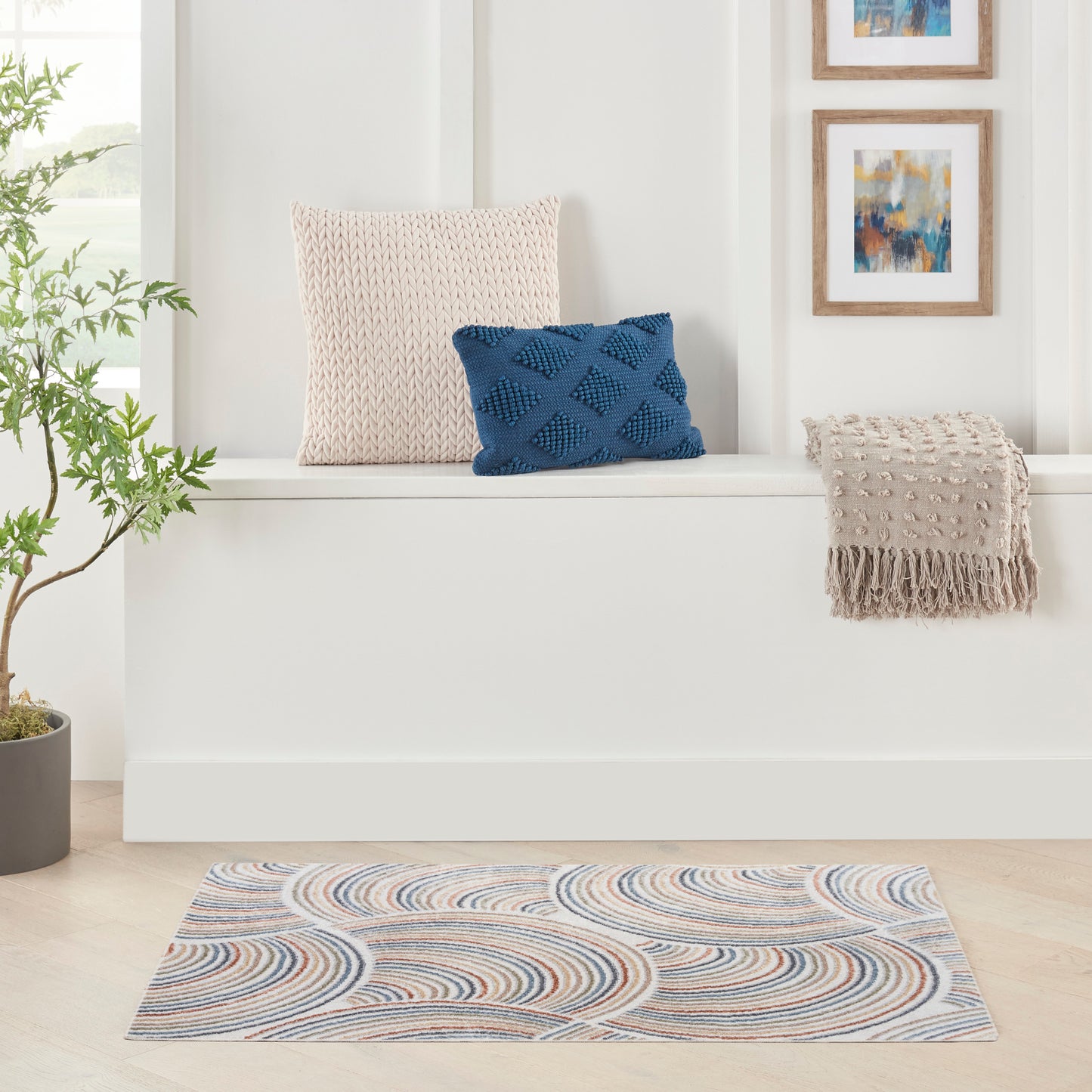 Beige knitted cushion and blue diamond pillow on a white window seat, with beige fringed throw and wavy blue-tan rug.