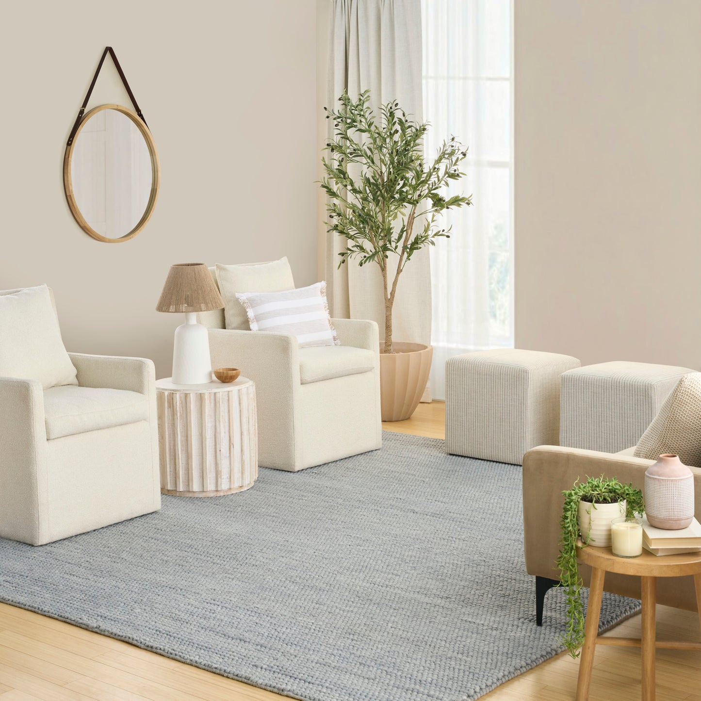 Cream armchairs in a beige living room with round wood table, tall plant, gray rug, circular wood-framed mirror.