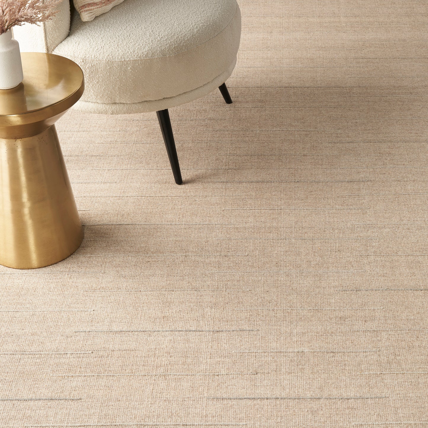 Beige round upholstered accent chair with black legs beside a gold metallic side table on a woven beige rug.