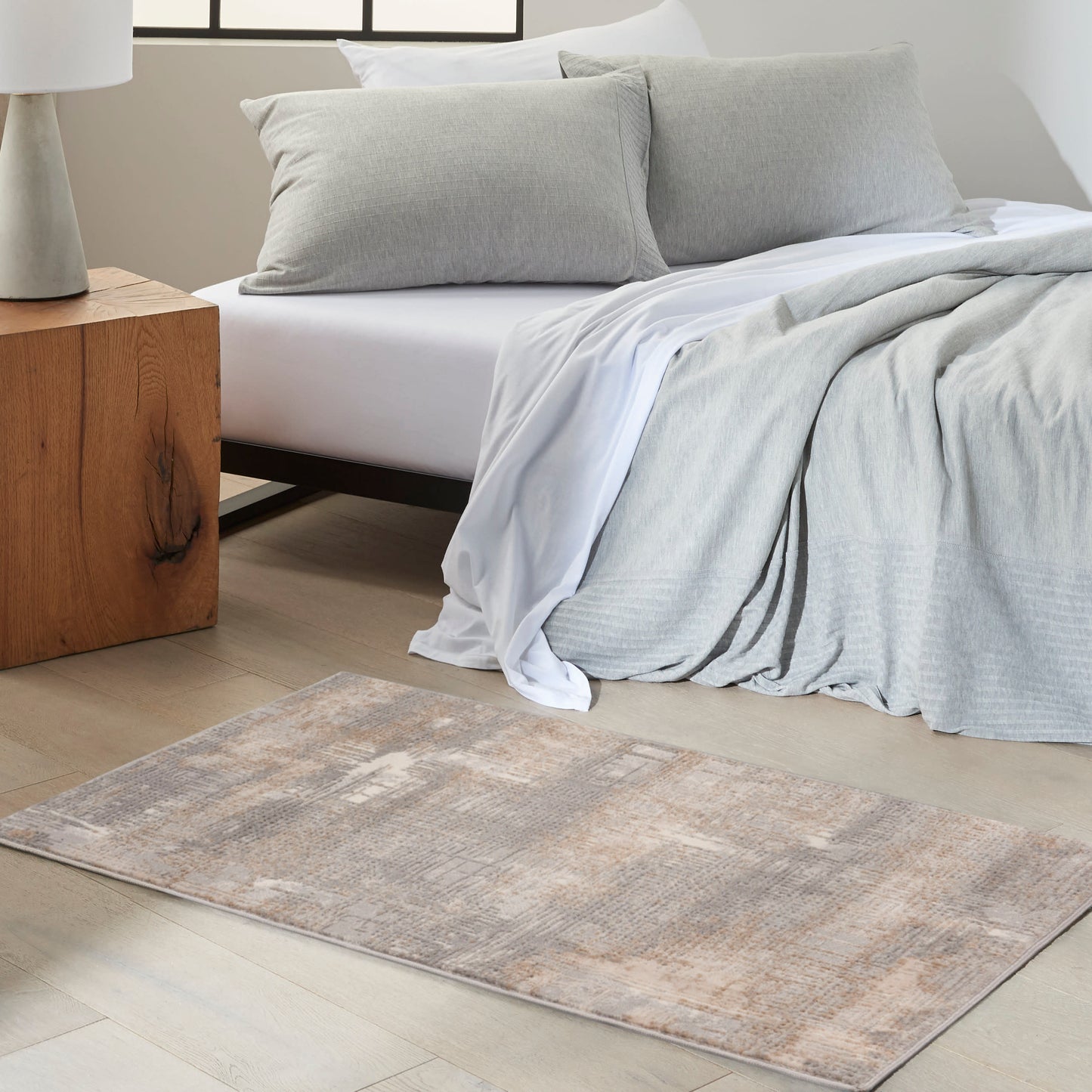 Neutral bedroom with light gray duvet, matching pillows, white sheets, beige-gray rug, and wooden block nightstand.
