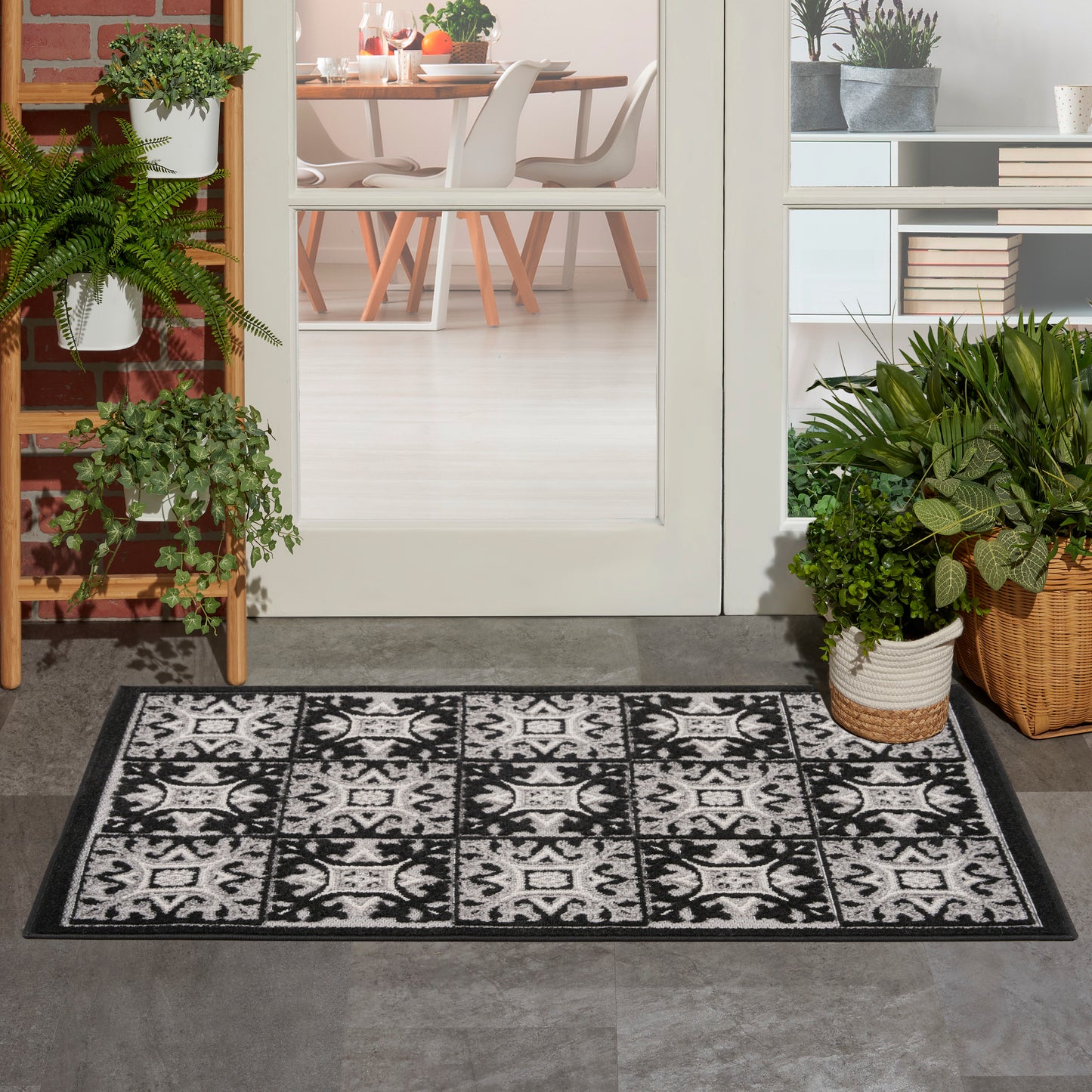 Rectangular black-and-white tile-patterned rug (3' x 4') on a gray floor, flanked by potted plants and glass doors to a dining room.