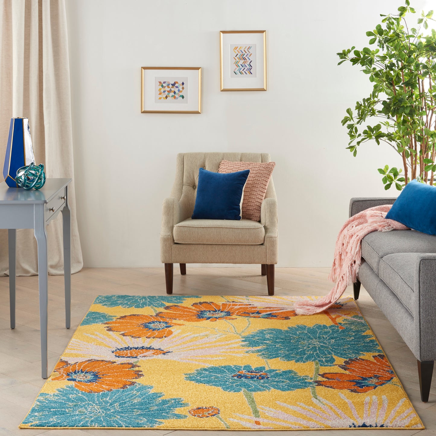 Cozy living room with a yellow, teal, and orange floral rug; beige armchair, gray sofa, blue cushions, pink throw, framed art.