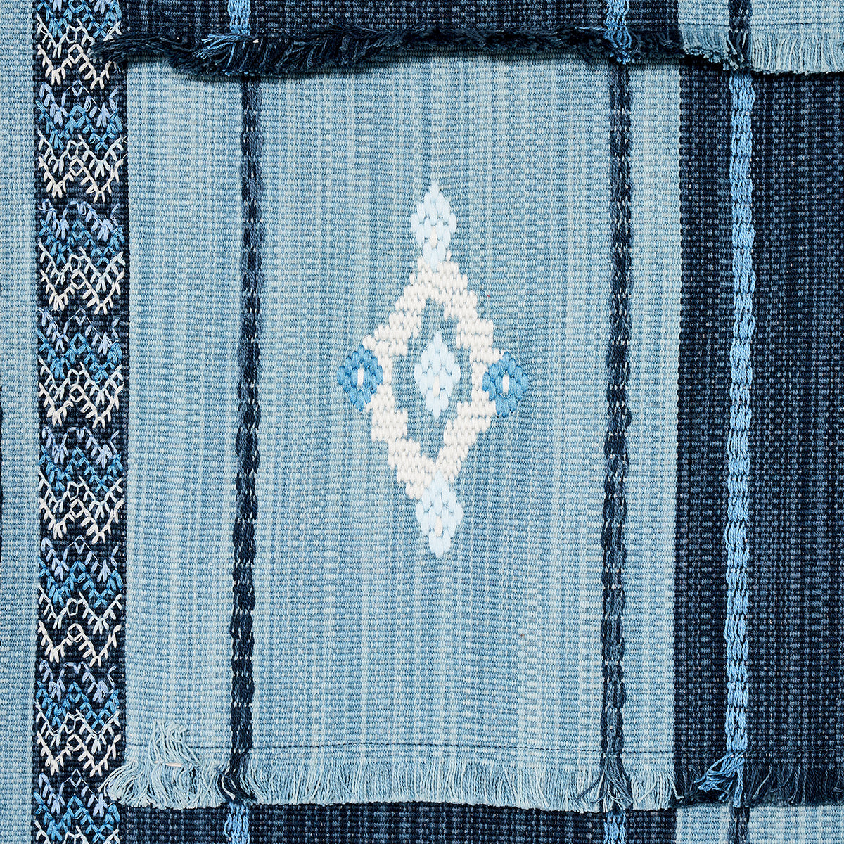 Blue woven rug with vertical stripes, central light-blue field with white diamond motif, dark blue borders and fringed edges.
