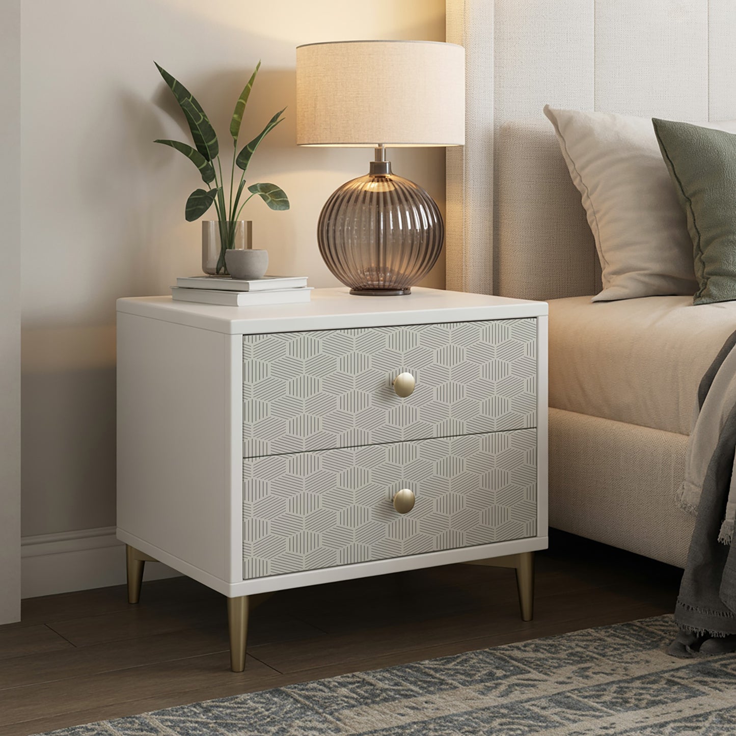 White two-drawer nightstand with geometric patterned fronts, brass knobs, tapered gold legs; lamp, plant, and books on top.