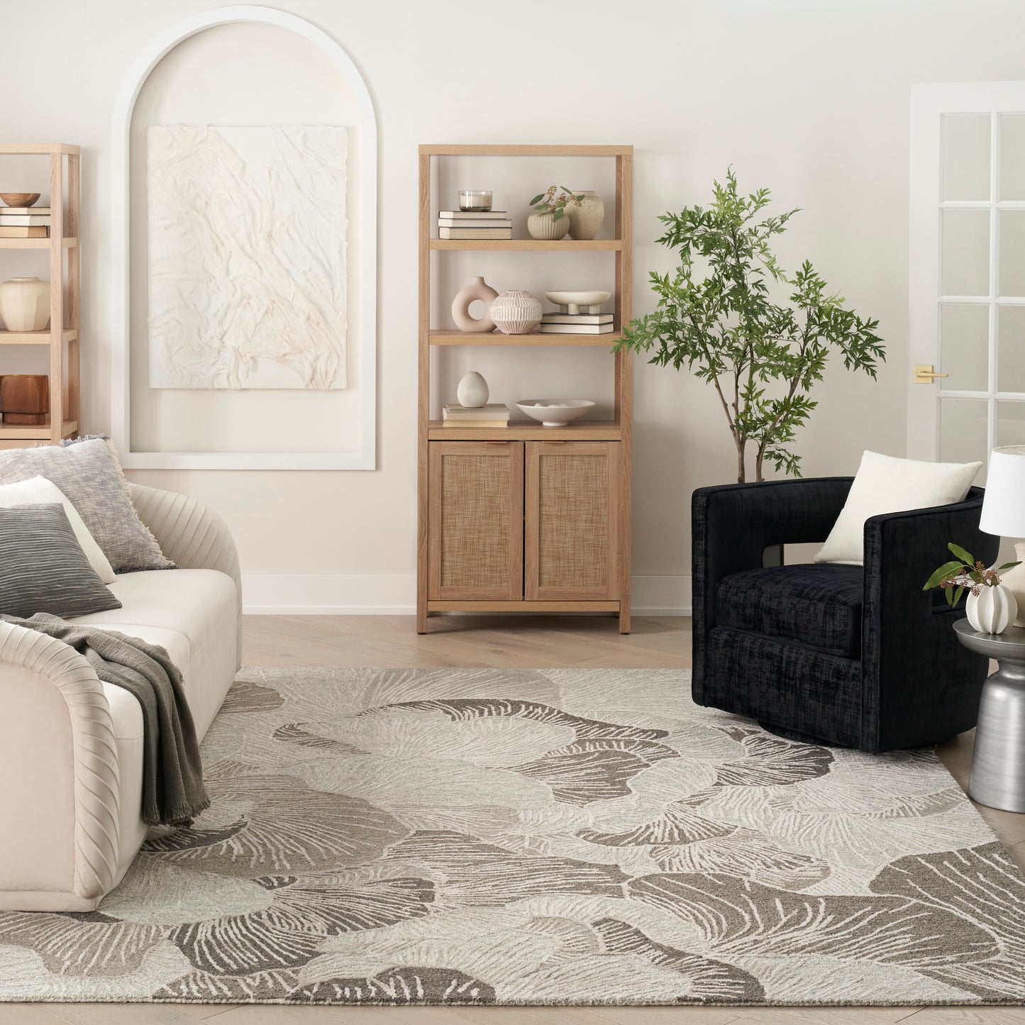 Neutral living room with beige sofa, black armchair, light wood shelving, potted plant, & a cream rug with leaf pattern.