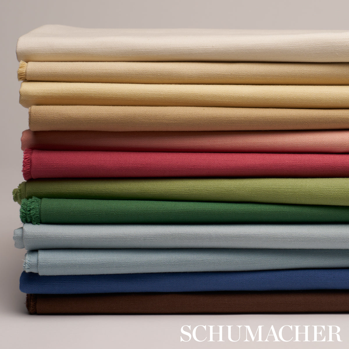 Stack of folded fabric in cream, beige, peach, pink-red, greens and blues; soft cotton-like textiles with woven texture.