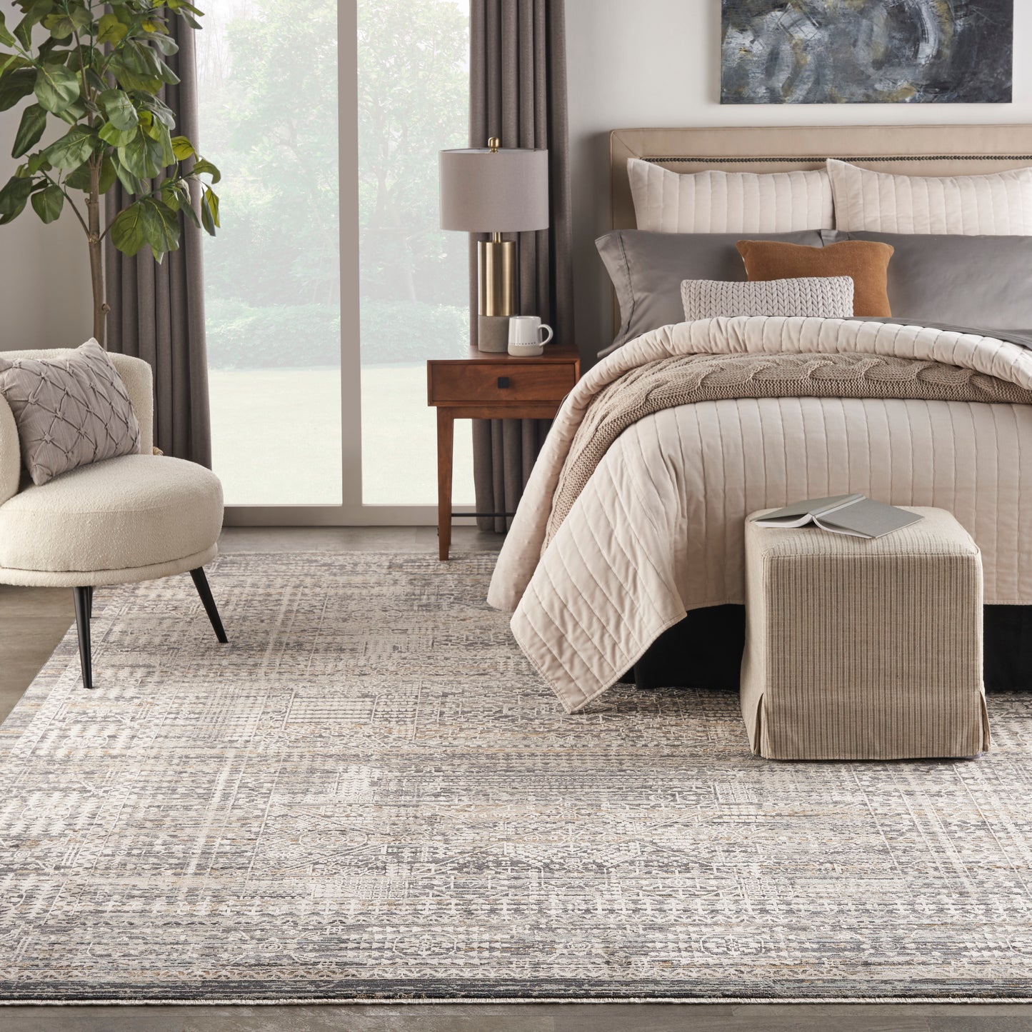 Neutral bedroom with tufted beige headboard, quilted cream bedspread, matching ottoman, wooden nightstand, brass lamp, plant, and patterned rug.