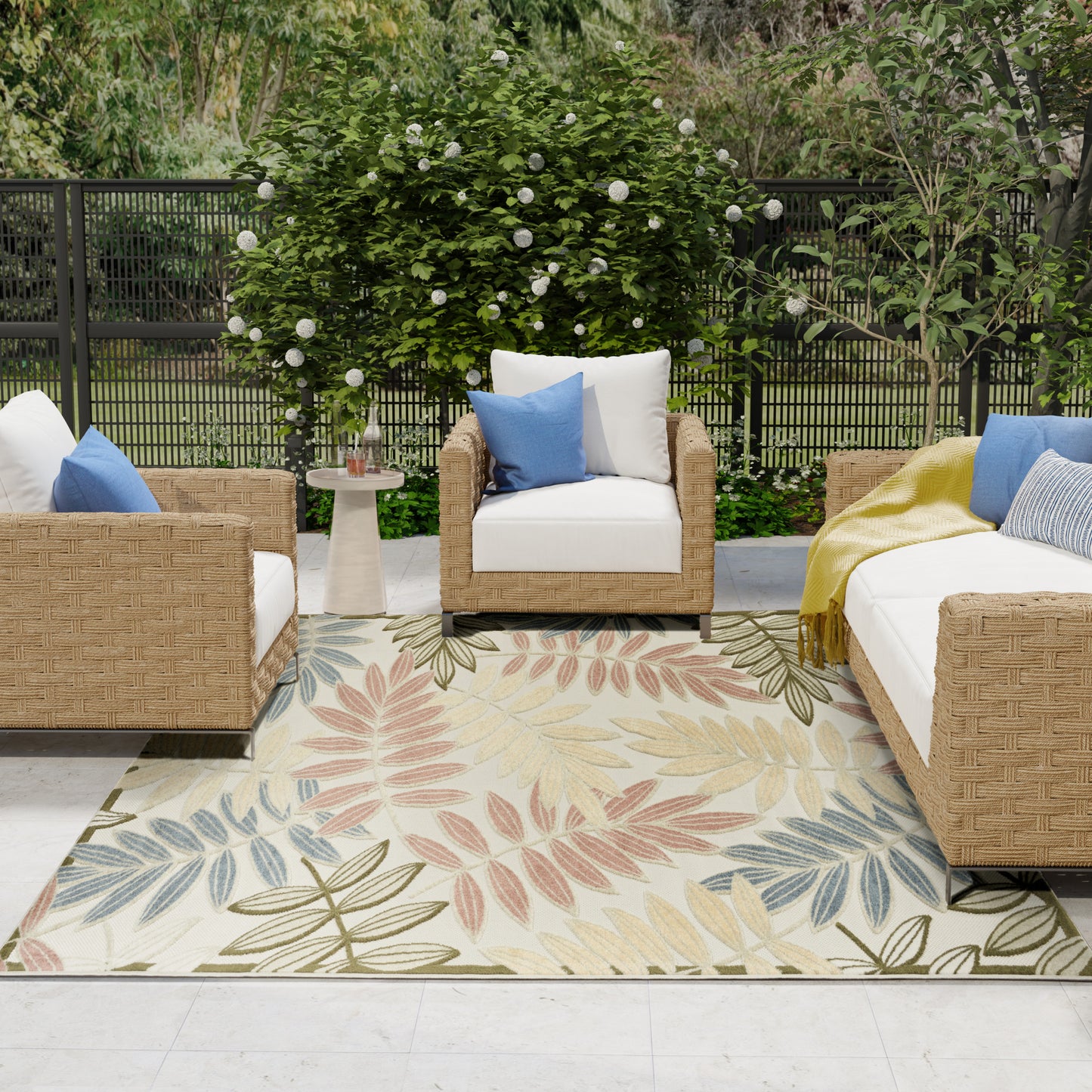 Outdoor wicker sofa and chairs with white cushions, blue pillows, a yellow throw, atop a pastel leaf-pattern rug in a garden.