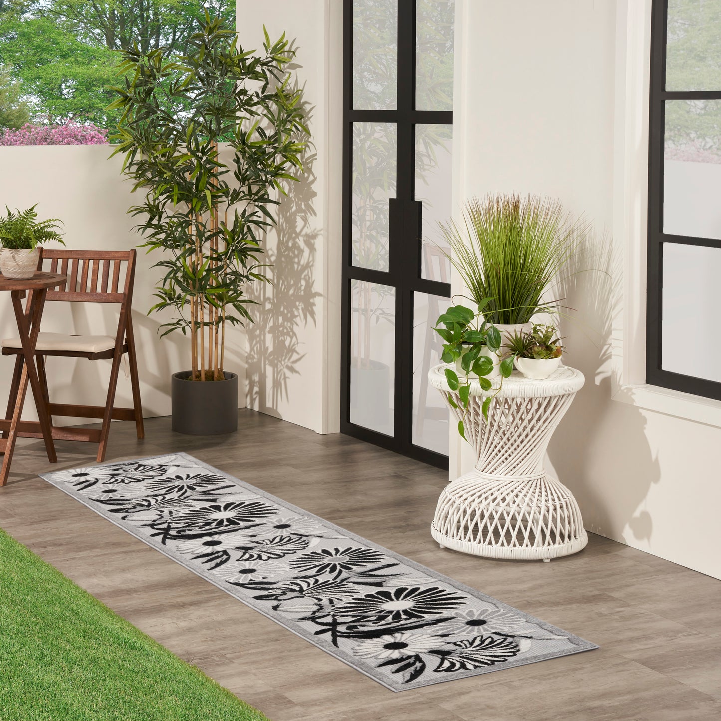 Long gray floral runner rug with black botanical pattern on wood floor beside glass doors and potted greenery.