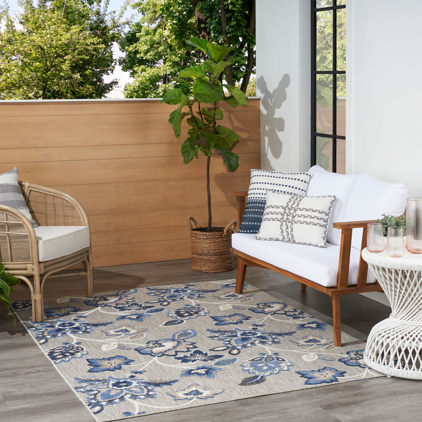 Outdoor balcony seating: white cushioned sofa with wood frame, rattan chair, blue floral rug, potted plant, wicker table.