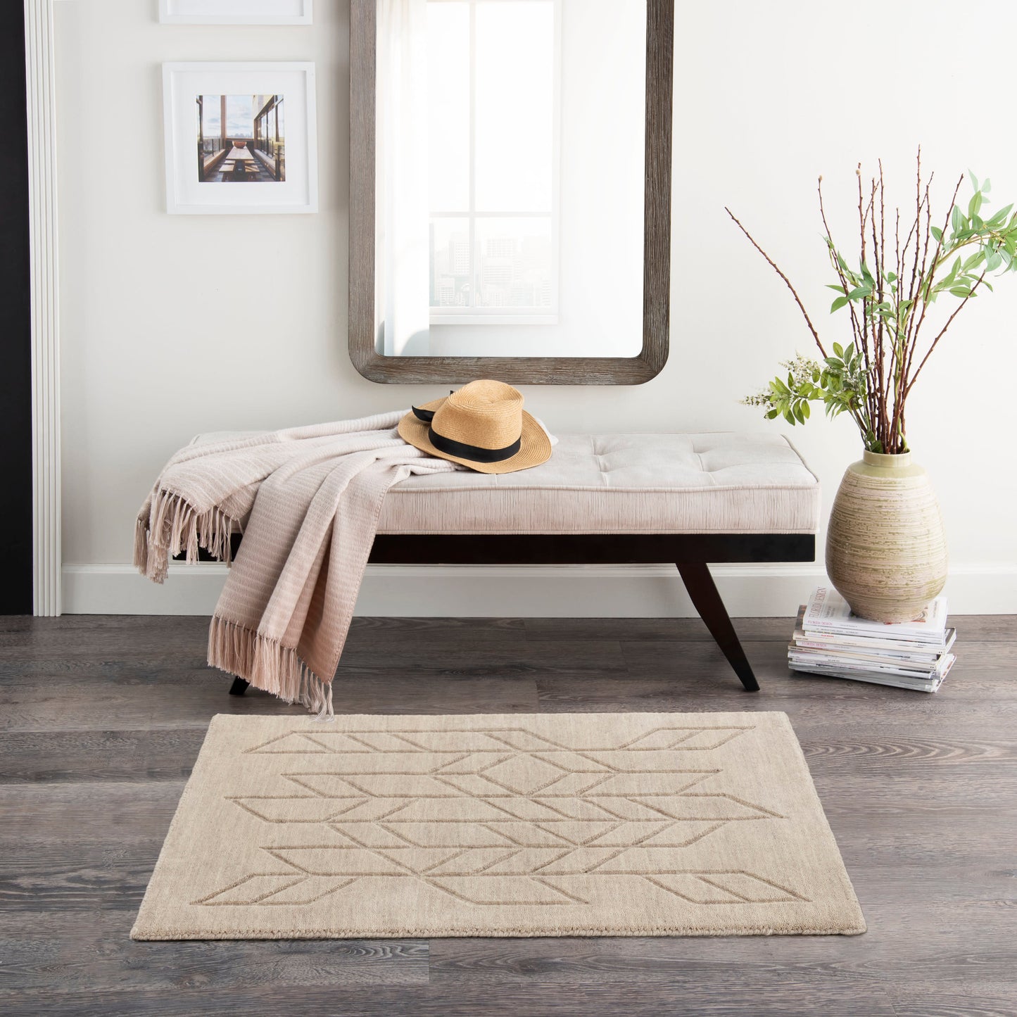 Beige upholstered bench with throw and straw hat, large dark wood-framed mirror, beige geometric rug, vase of branches.
